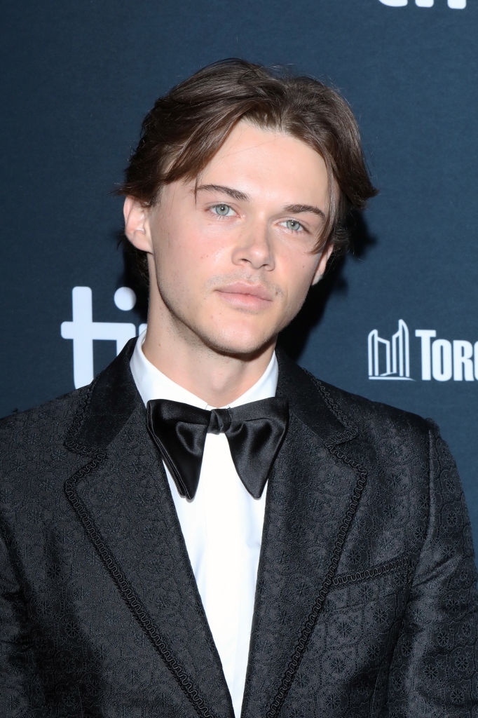 Person successful  ceremonial  attire with a acheronian  textured suit   and bow necktie  astatine  a ceremonial  event; backdrop features logo pattern