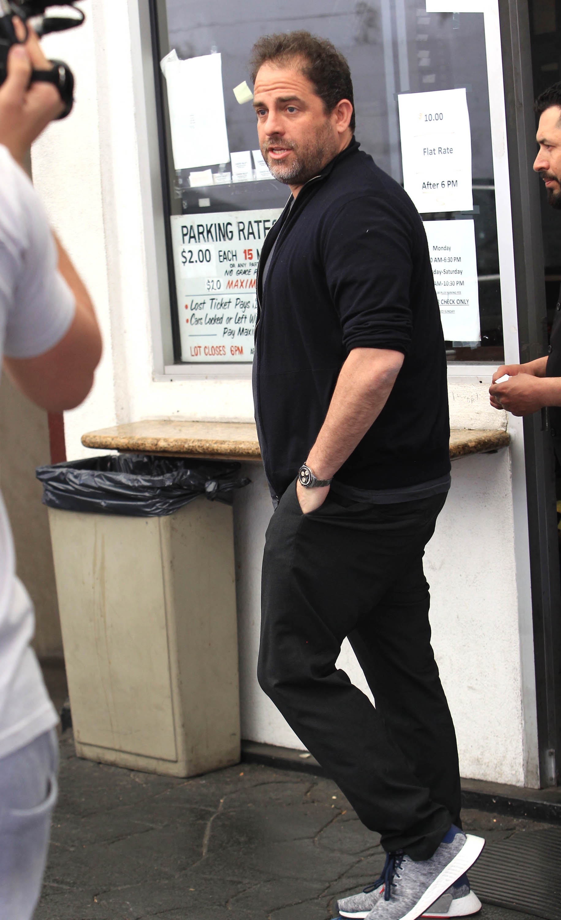 Brett Ratner i uformelt antrekk, inkludert en jakke og joggesko, står på et fortau, med et parkeringsskilt synlig i bakgrunnen