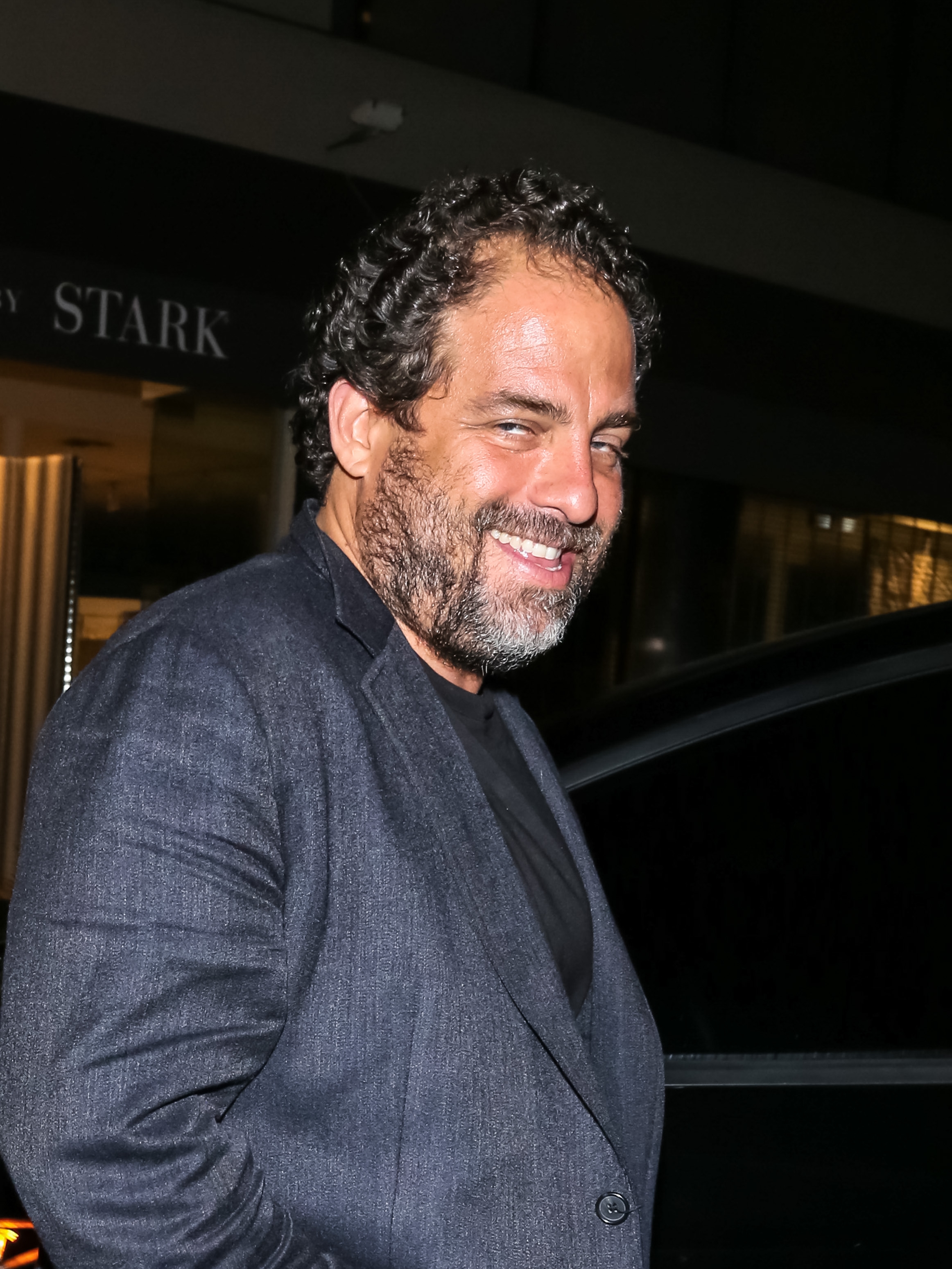 Person smiling, wearing a dark blazer over a t-shirt, standing outside a building