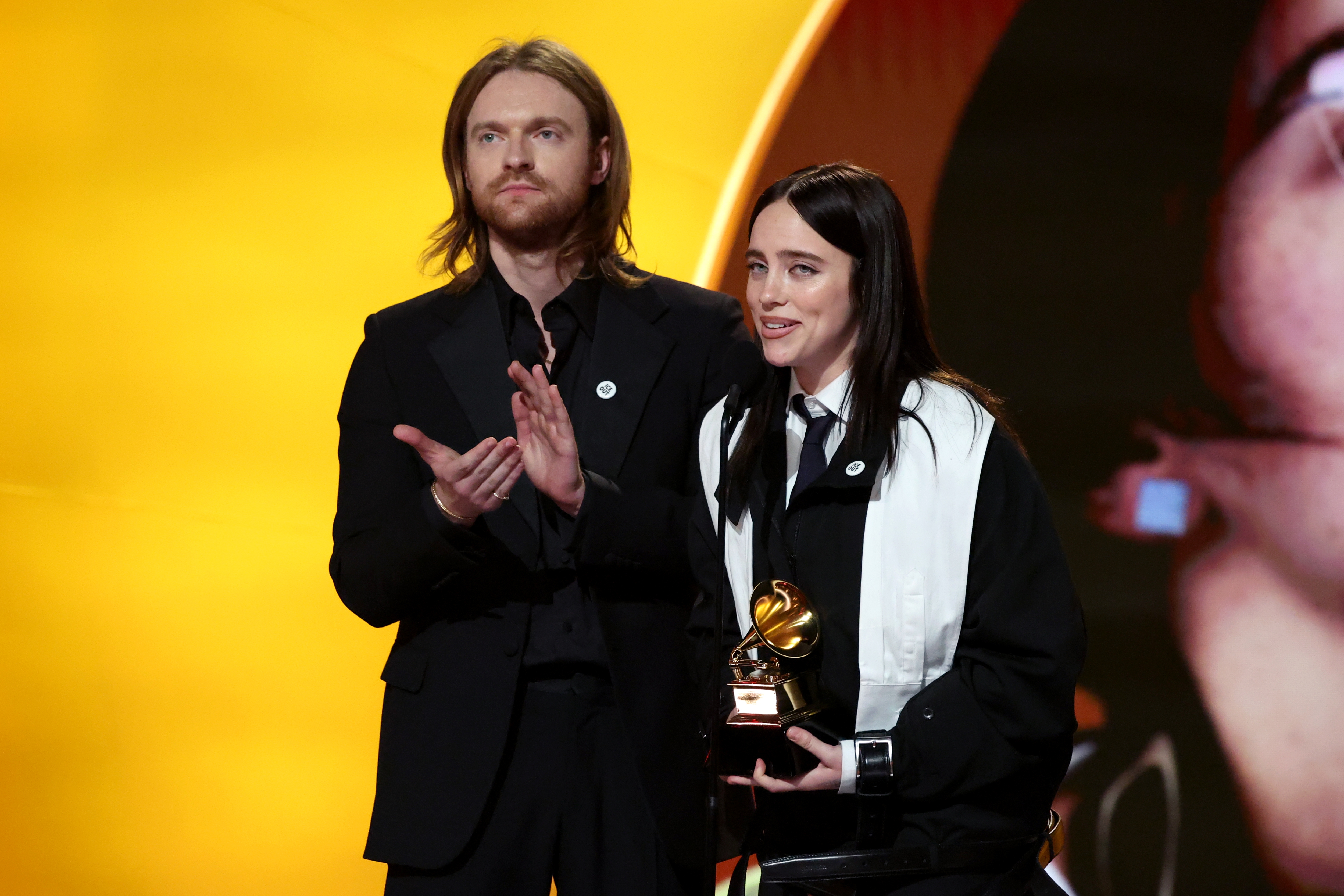 Two radical   basal   connected  signifier    with a Grammy award. One wears a suit, the different   a achromatic  outfit with a achromatic  scarf
