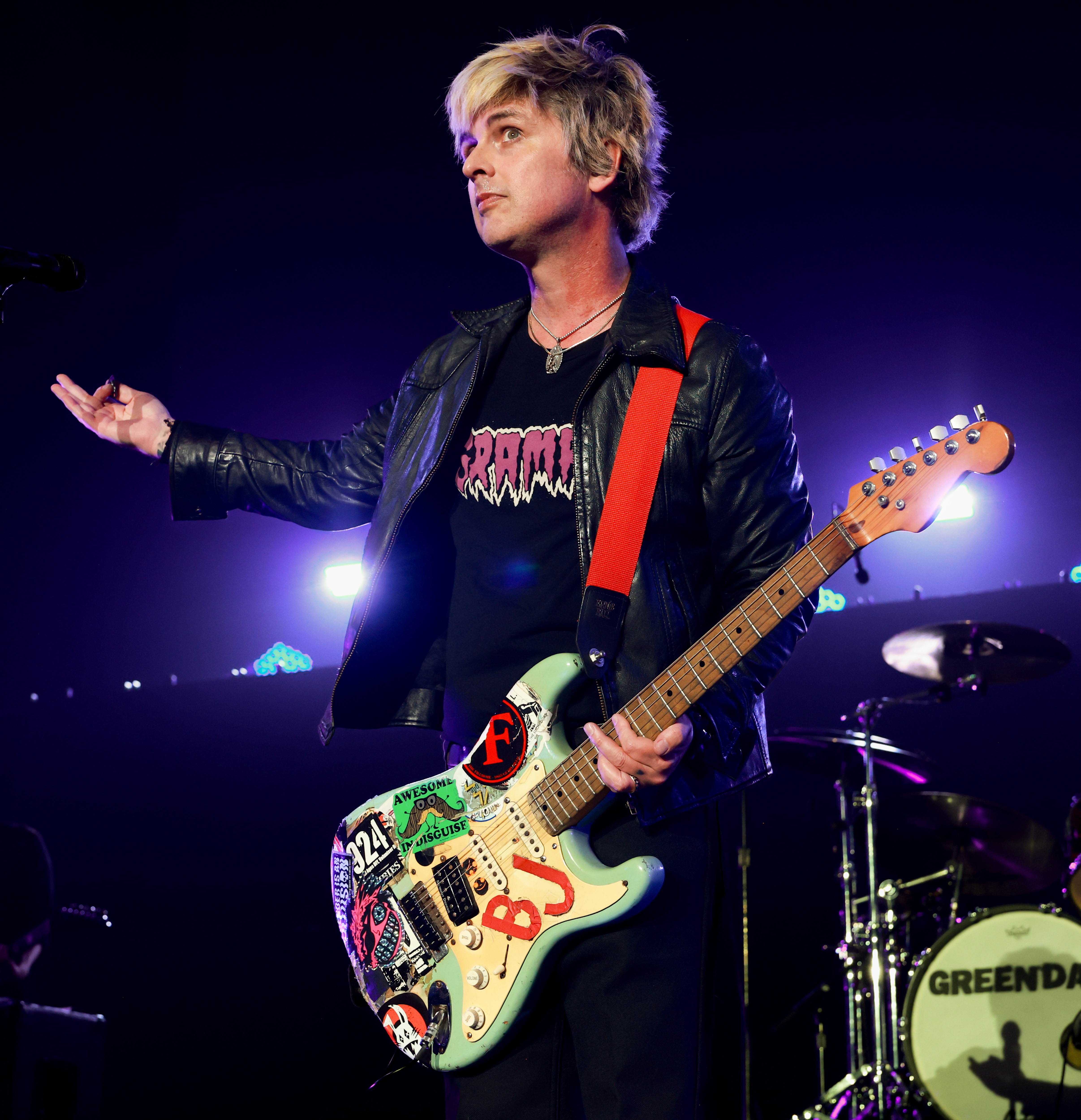 Musician onstage with electric guitar decorated with stickers, wearing a black leather jacket and graphic tee, performing energetically