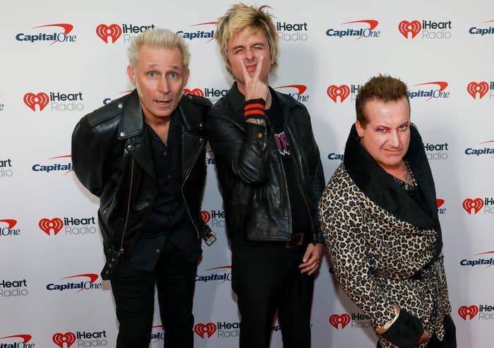 Three individuals pose playfully on a red carpet at an iHeartRadio event. Two wear leather jackets; one wears a patterned coat
