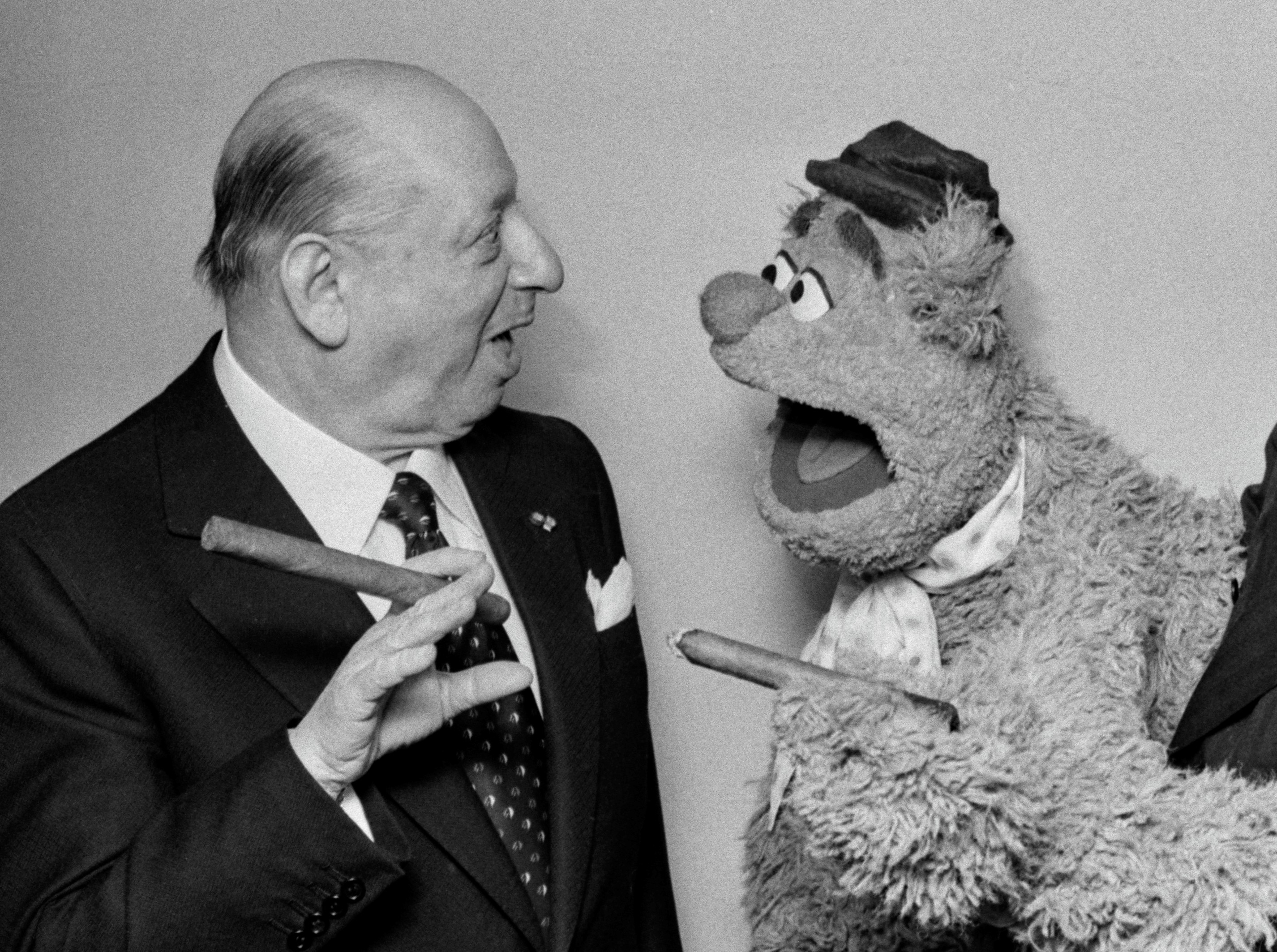 Lord Lew Grade successful a suit and necktie holds a cigar, chatting with Fozzie Bear dressed successful a chapeau and scarf, who is besides holding a cigar