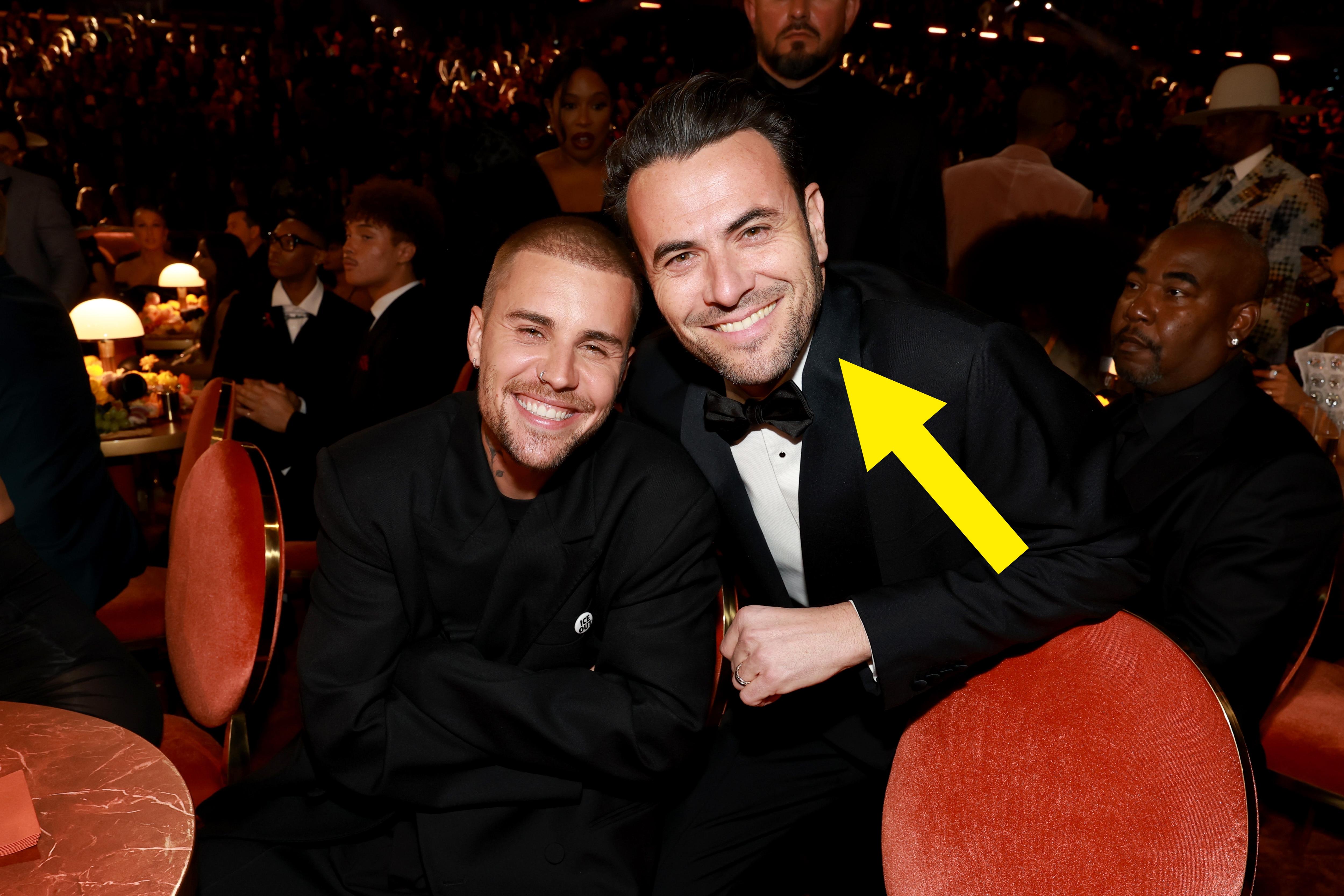 Two radical   smiling, seated astatine  an event. One wears a suit, the different   successful  a casual outfit. Surrounded by a crowded, elegantly dressed audience