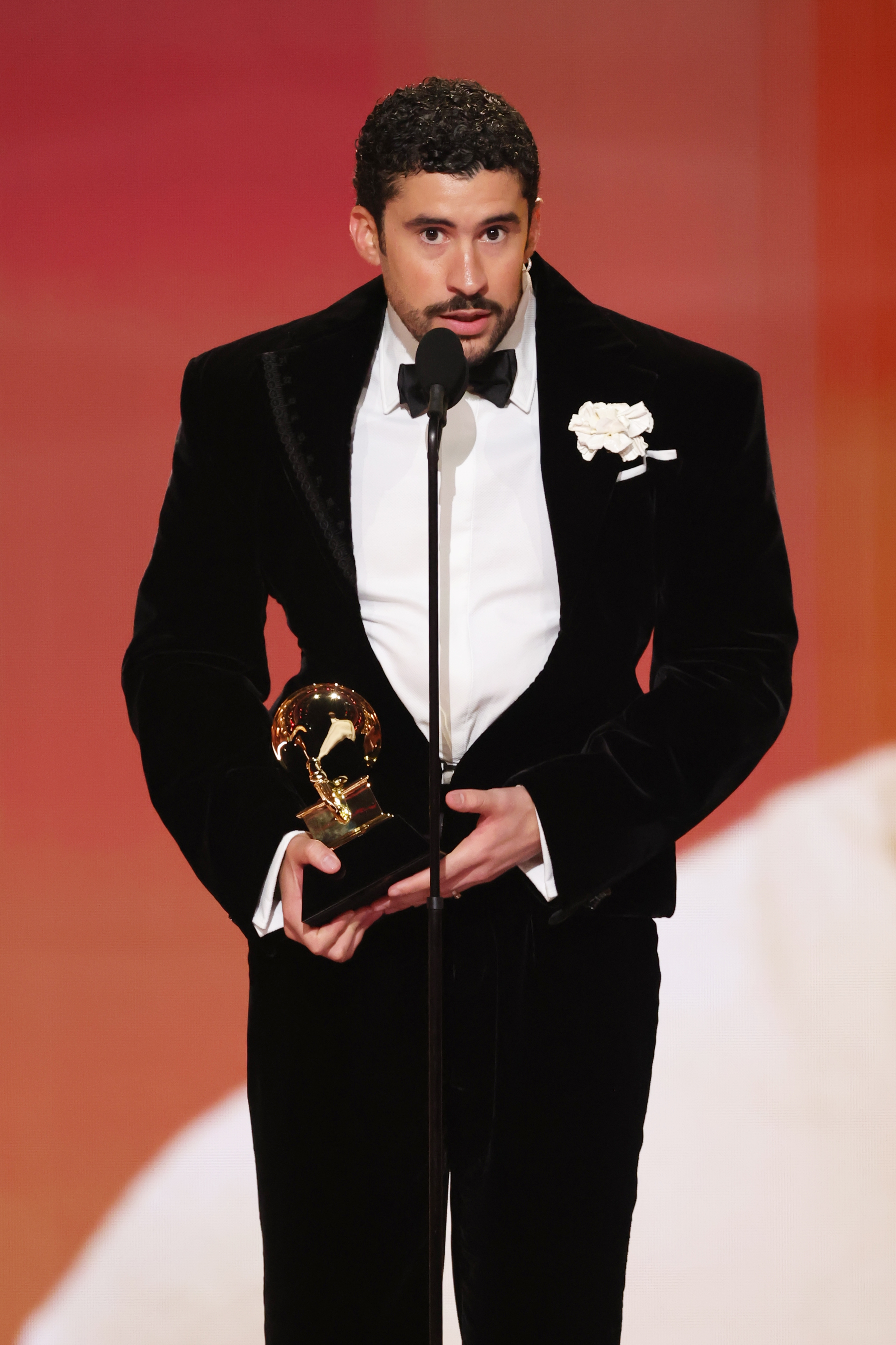Person successful  elegant achromatic  velvet suit   with achromatic  garment  and angiosperm  connected  lapel, holding an award, speaks astatine  a podium astatine  a ceremonial  event