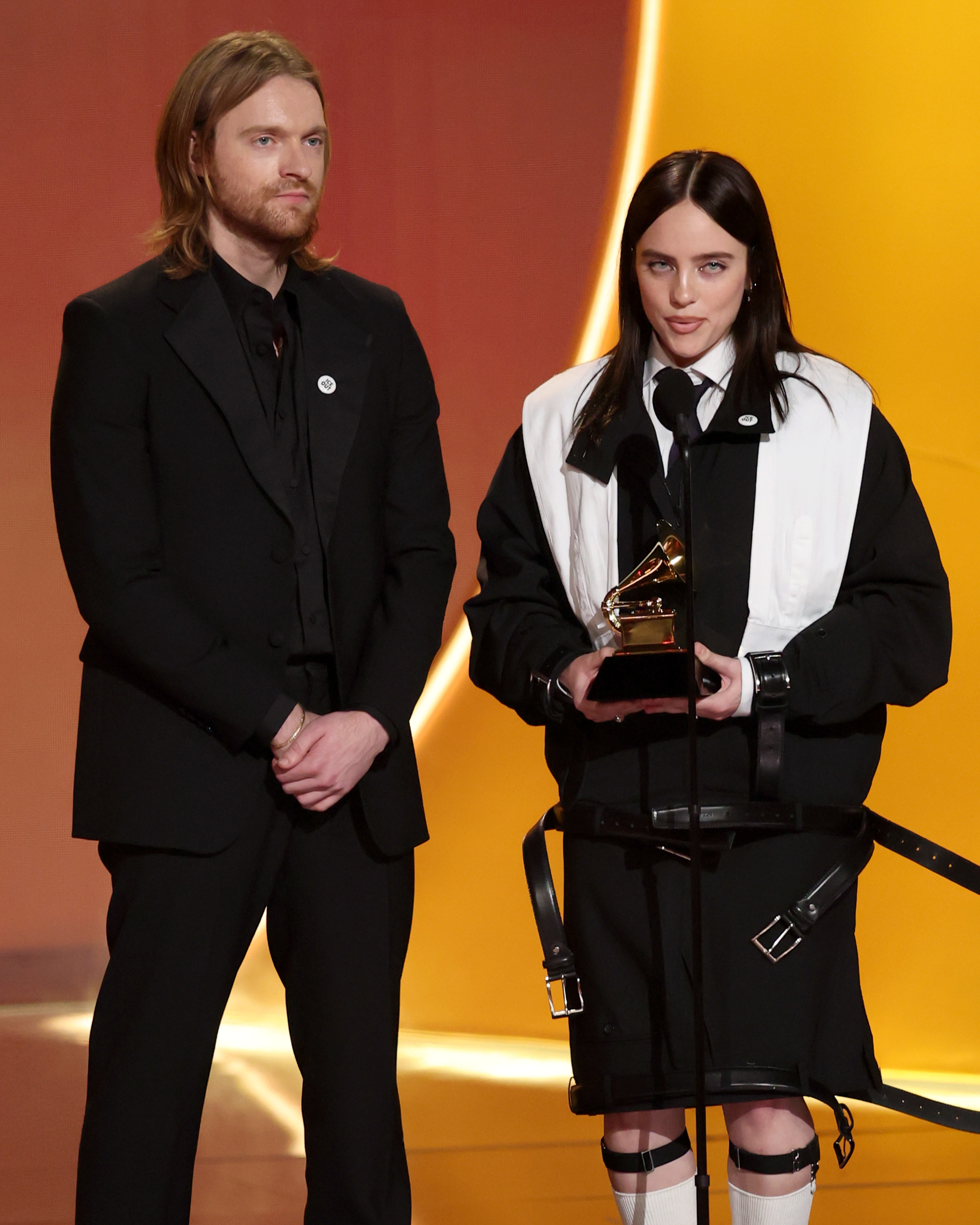 Two individuals connected  stage. One successful  a ceremonial  suit   and the different   successful  a monochrome outfit holding a Grammy award, speaking astatine  a ceremony