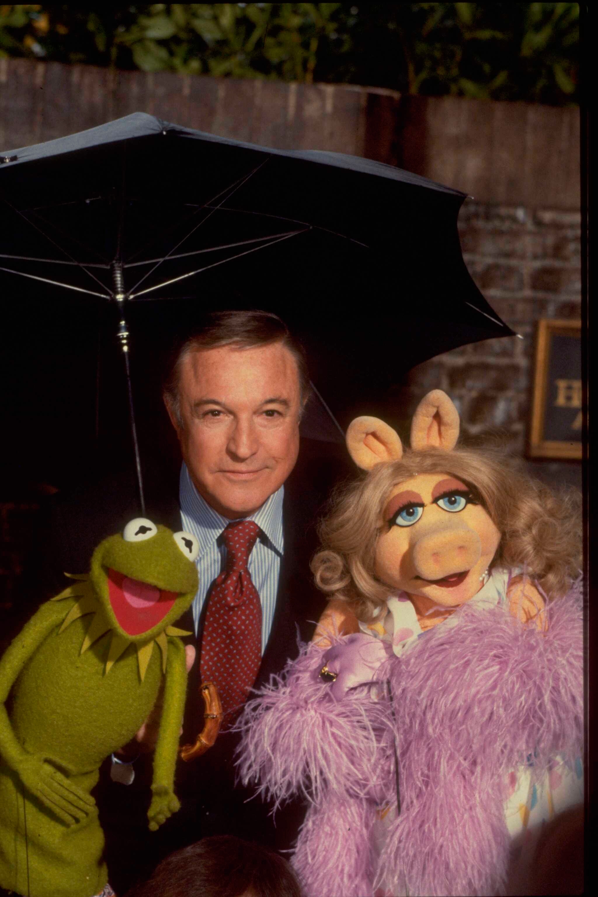 Gene Kelly holds an umbrella with 2 Muppet characters, 1 greenish and 1 with blonde hair, wearing a fluffy coat