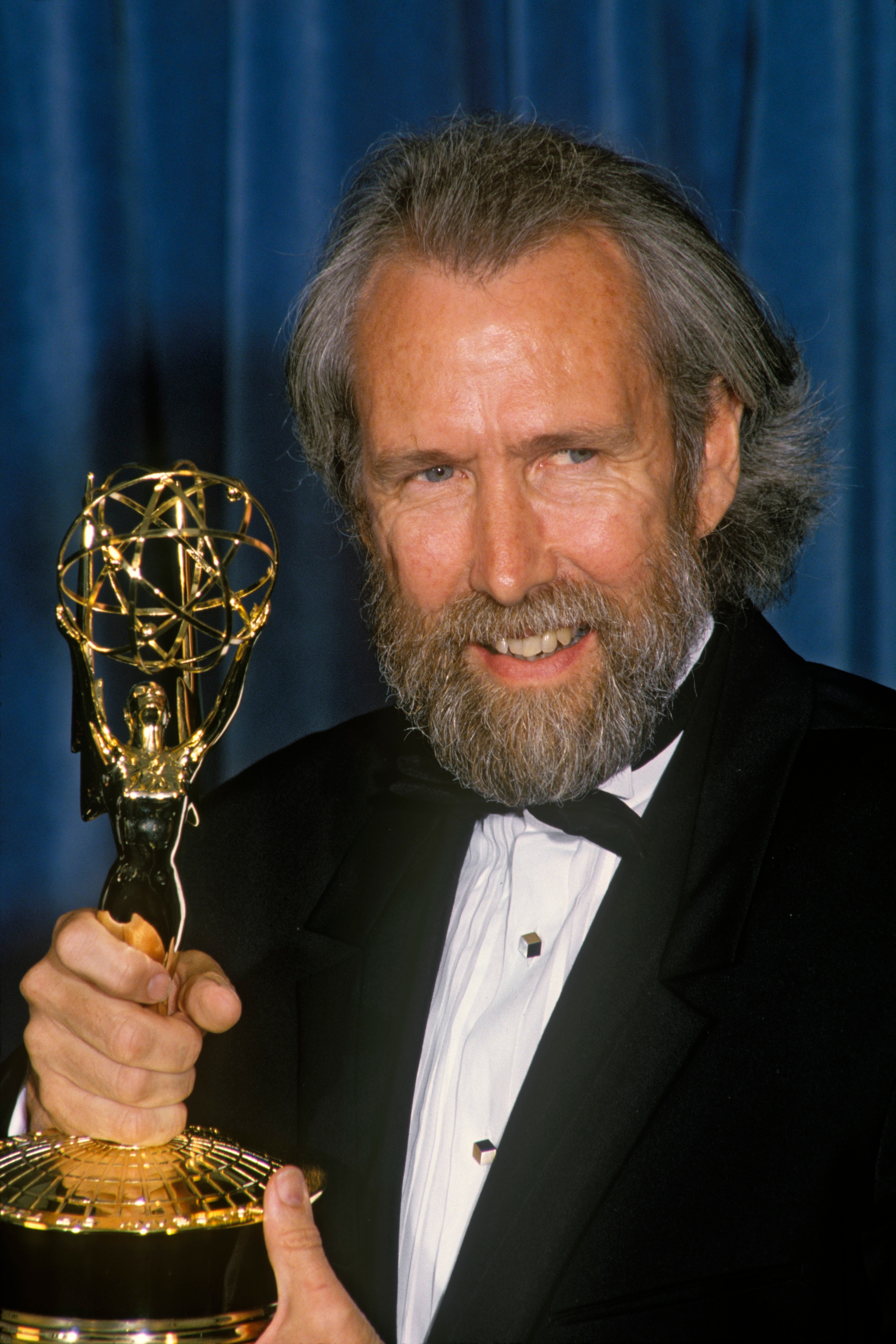 Jim Henson successful a tuxedo holding an Emmy award, smiling successful beforehand of a bluish backdrop