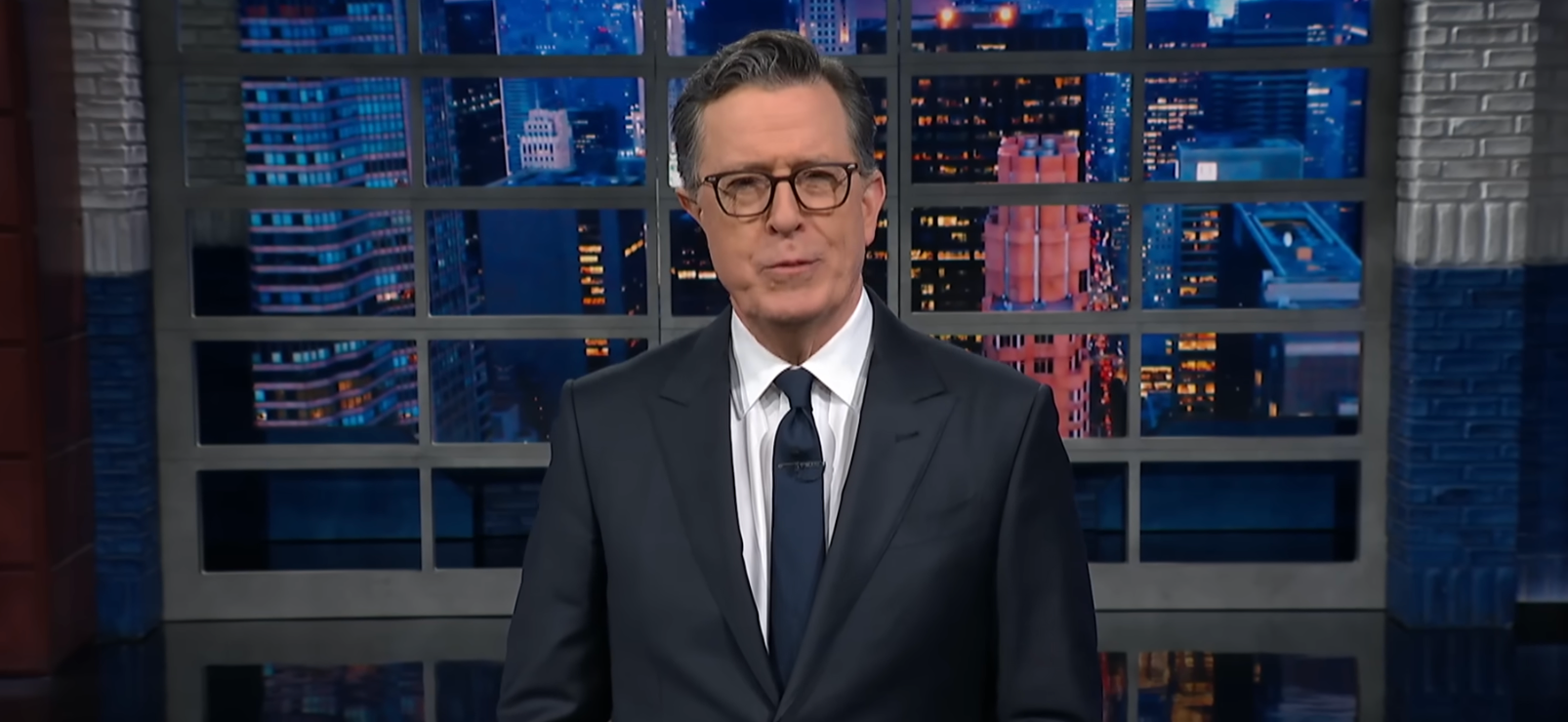 En person i dress og slips står på et talkshowsett med bybildebakgrunn