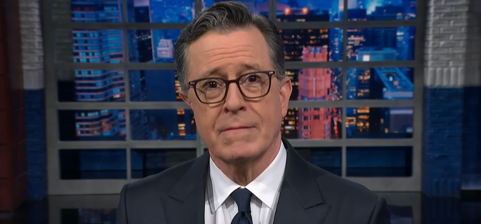 En person med briller, iført dress og slips, vises på et TV-apparat med et bybilde i bakgrunnen