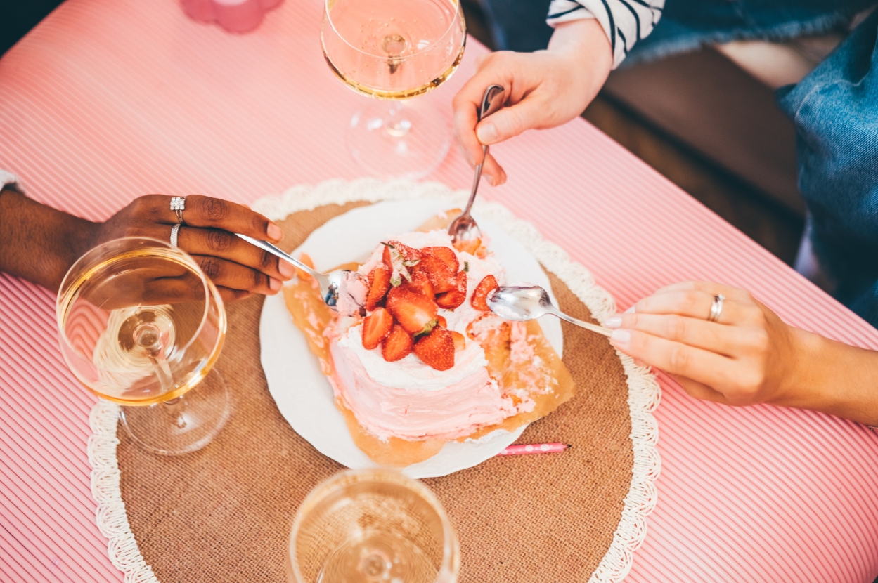 Three people enjoying a strawberry-topped dessert at a table with wine glasses, sharing with spoons