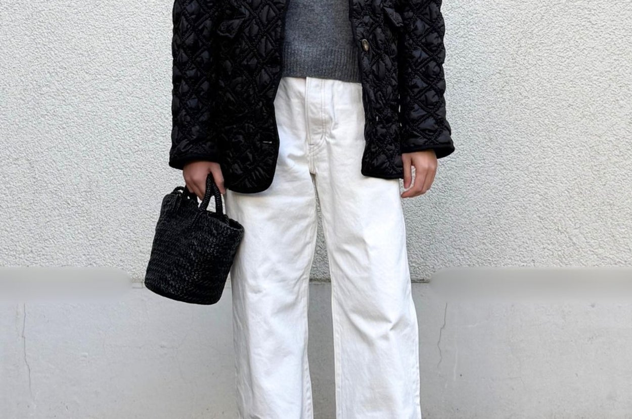 女性が模様入りのジャケット、グレーのセーター、白いワイドパンツを着用し、手に黒いバッグを持っている。
