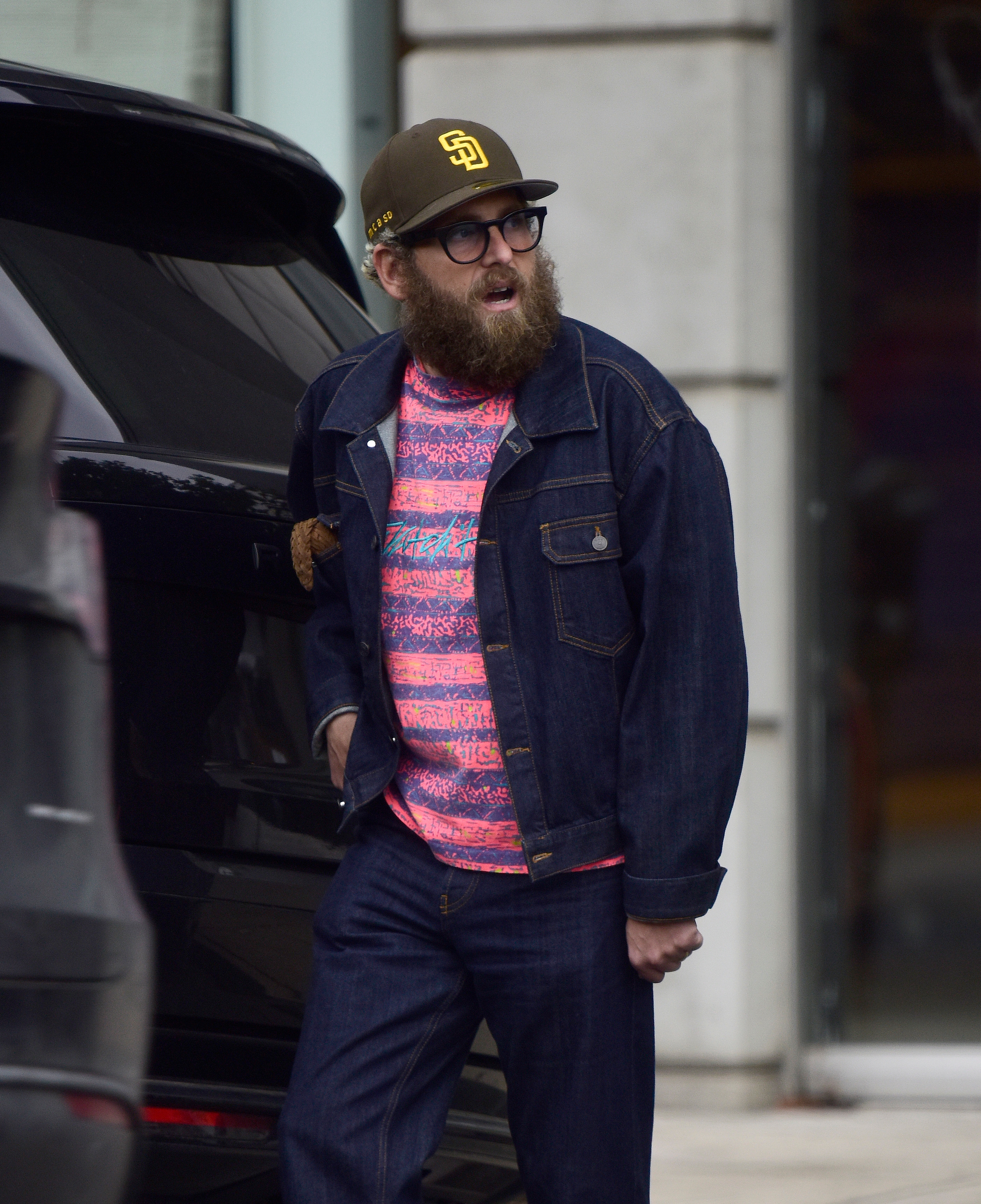 A bearded idiosyncratic  successful  glasses wears a patterned shirt, denim jacket, and pants, with a shot   cap, lasting  by a conveyance  connected  a metropolis  street