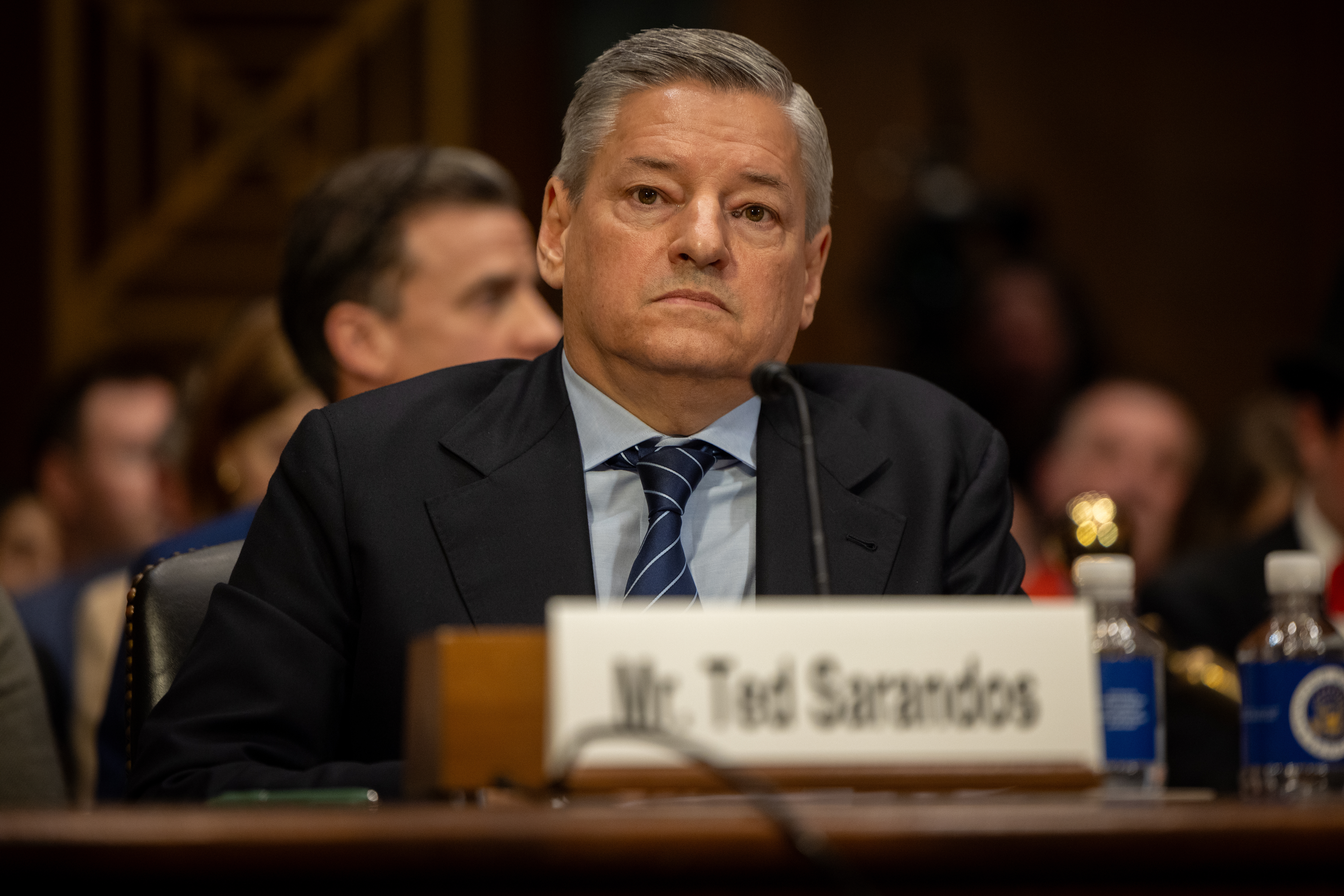 Jeg vet ikke hvem denne personen er, men de sitter ved et bord i formelle omgivelser, iført dress og slips, med navneskilt foran seg