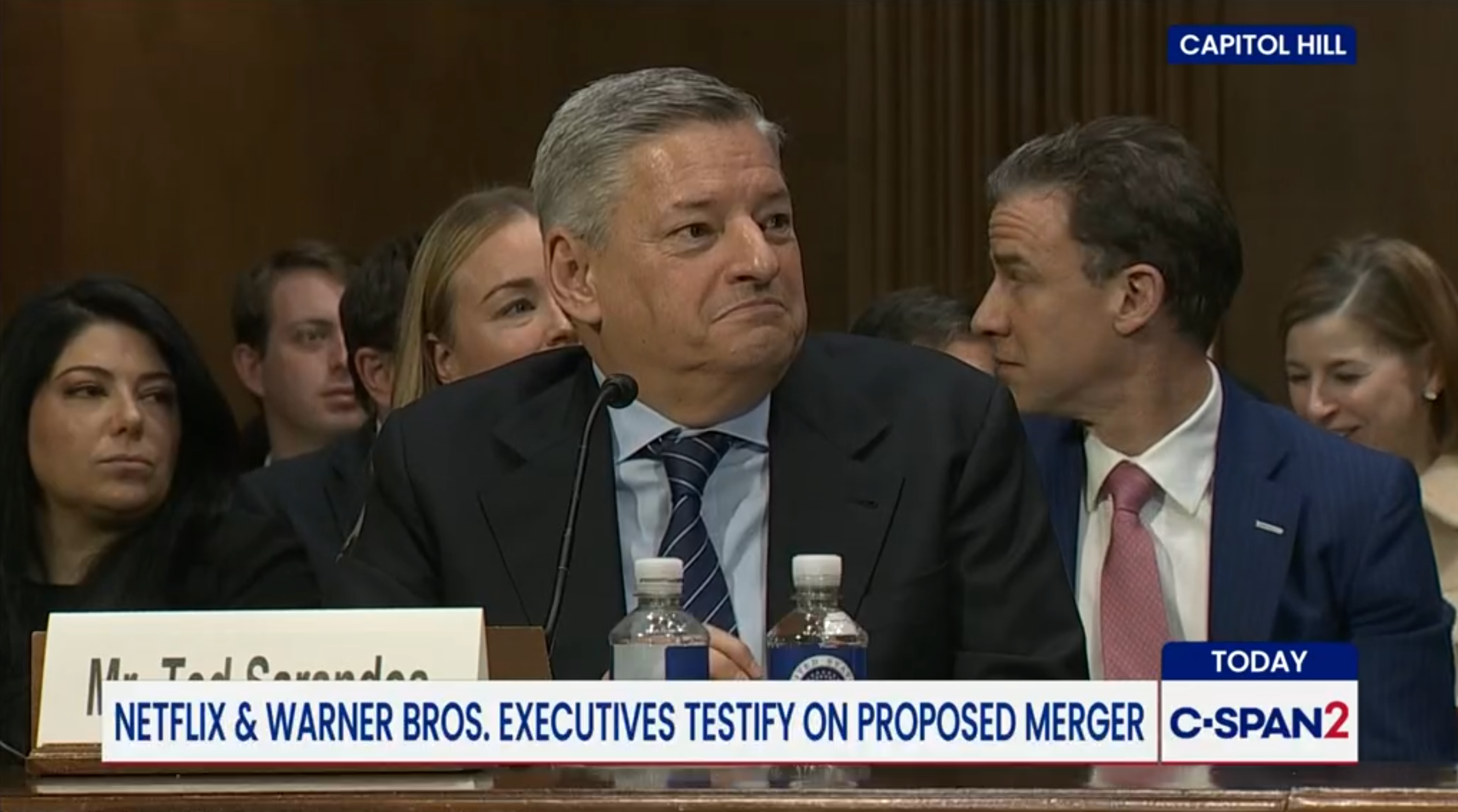 A man, perchance  an executive, is sitting astatine  a proceeding  with a nameplate. People are seated down  him, indicating a ceremonial  setting