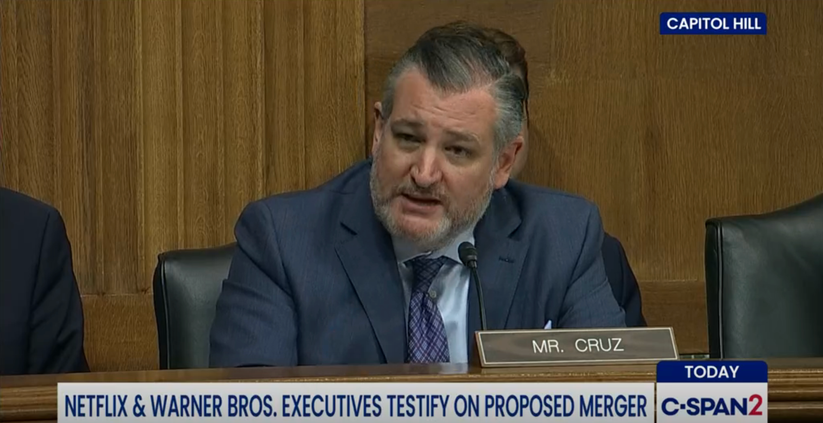 En mann i dress taler under en høring i Capitol Hill. Skjermteksten indikerer en diskusjon om en sammenslåing av Netflix og Warner Bros