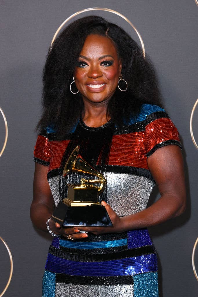 A idiosyncratic   successful  a sequined, geometric-patterned formal  holds a Grammy award, smiling connected  the event's backdrop