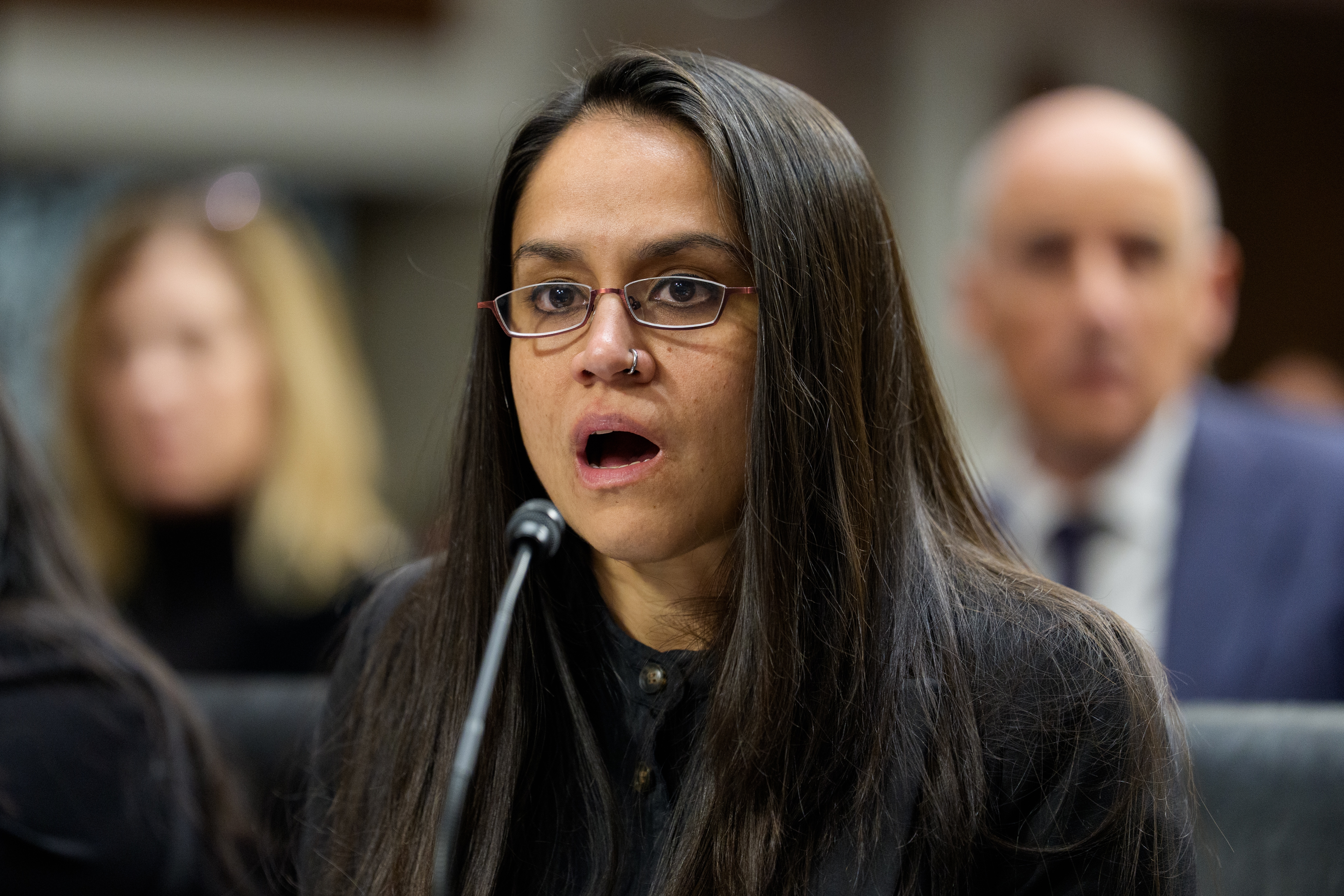 En person med langt hår og briller snakker ved en mikrofon under en offentlig høring, med oppmerksomme personer i bakgrunnen