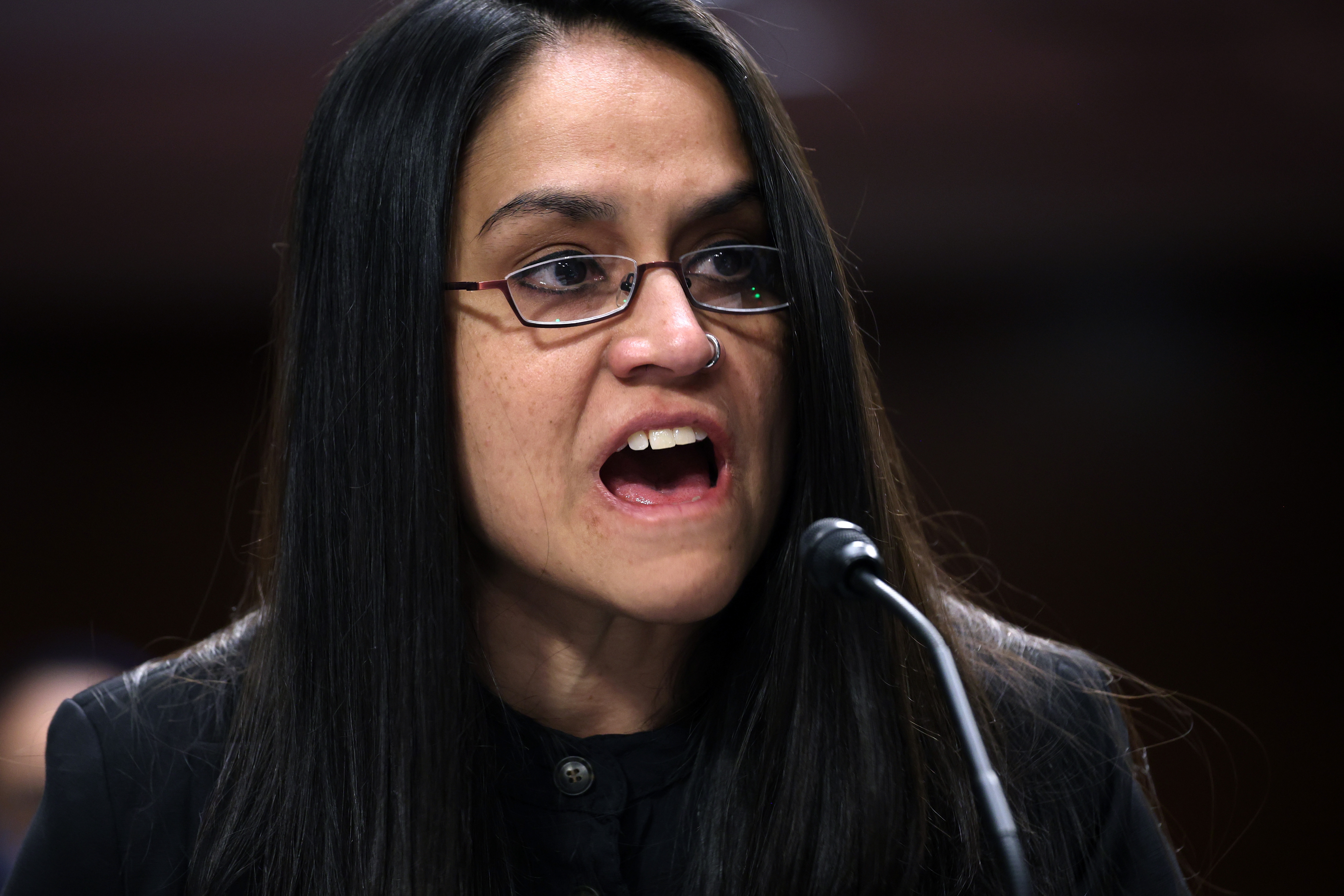 Person som snakker lidenskapelig inn i en mikrofon på et offentlig eller formelt arrangement, iført briller og mørkt antrekk