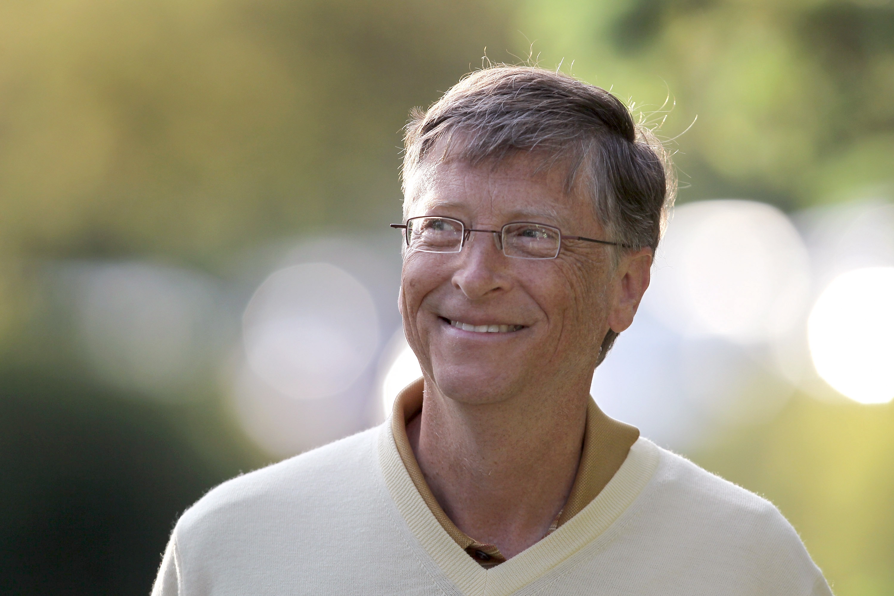 A antheral   with glasses and a sweater smiles outdoors, surrounded by a softly blurred background