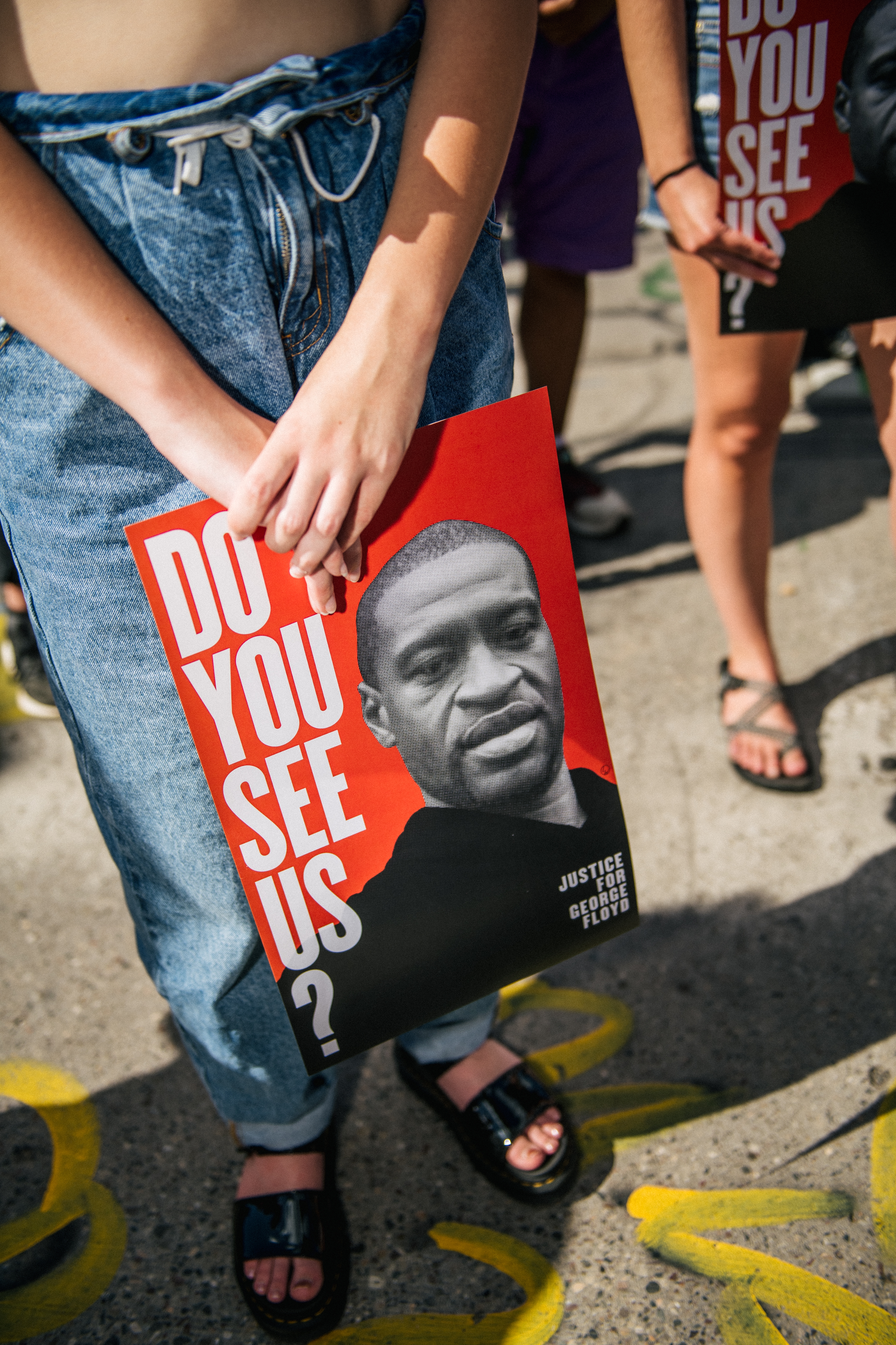 Person som holder en "Ser du oss?" plakat med George Floyds bilde under en protest. Teksten inkluderer "Justice for George Floyd."