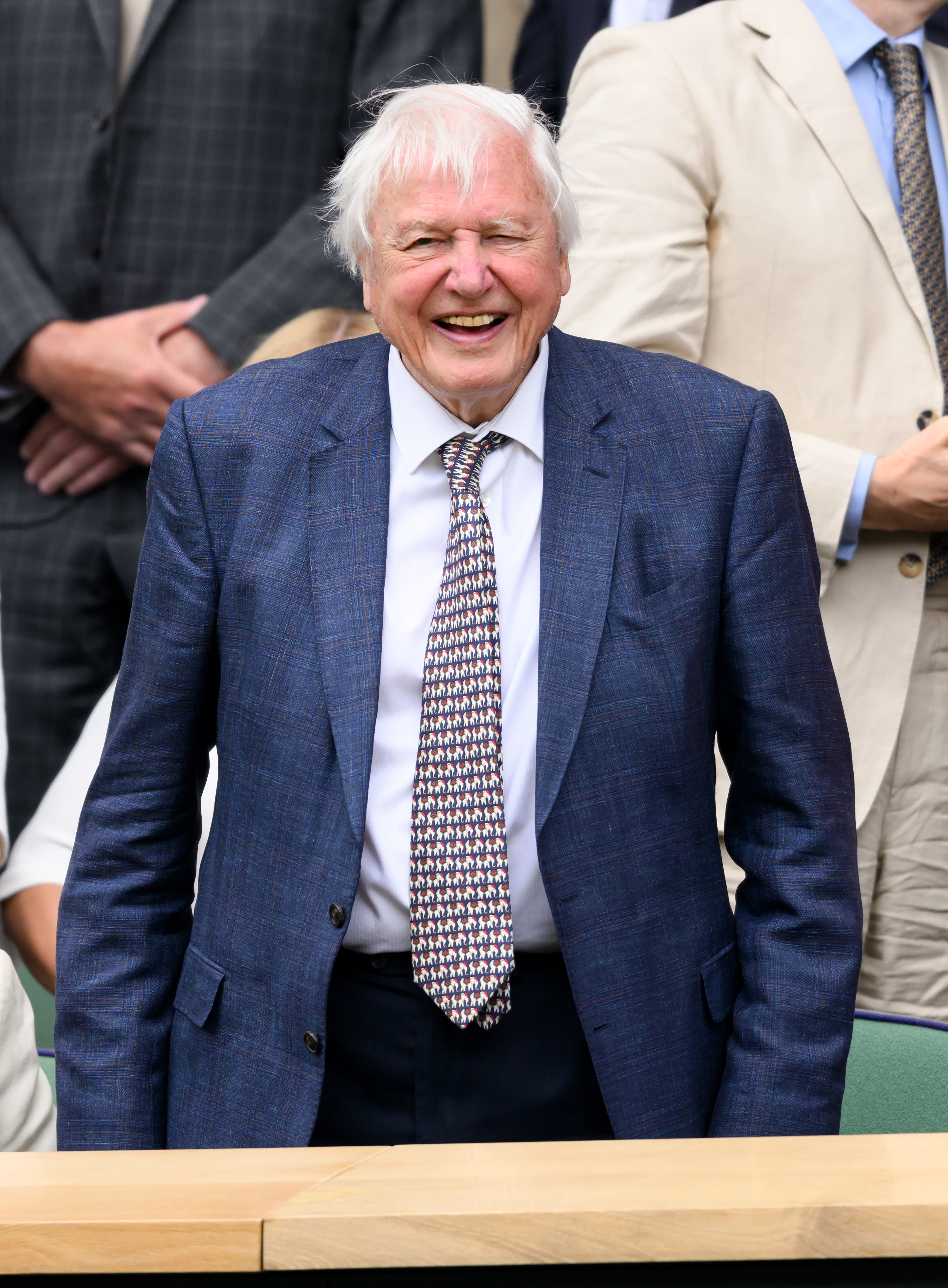 An older man in a suit and patterned tie smiles while attending an outdoor event, surrounded by others