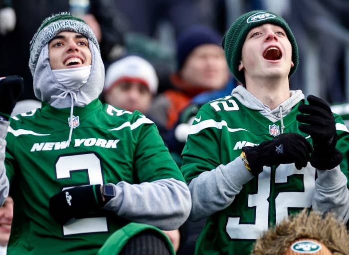 Two enthusiastic football fans in matching New York jerseys cheer during a game. They're wearing winter hats and gloves, bundled up against the cold