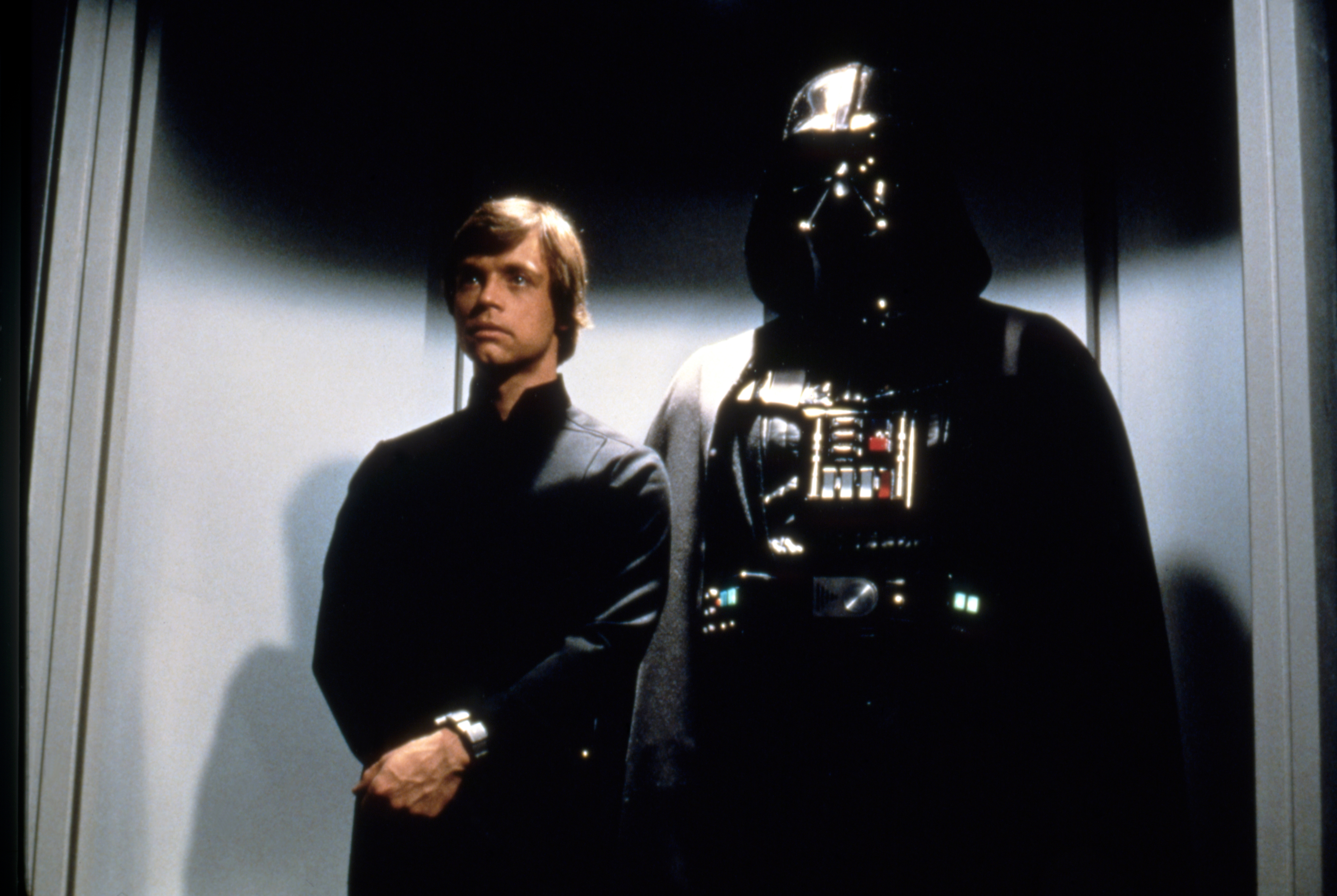 Two iconic sci-fi characters stand side by side in a futuristic setting, one in a dark suit and helmet, and the other in a simpler, dark outfit