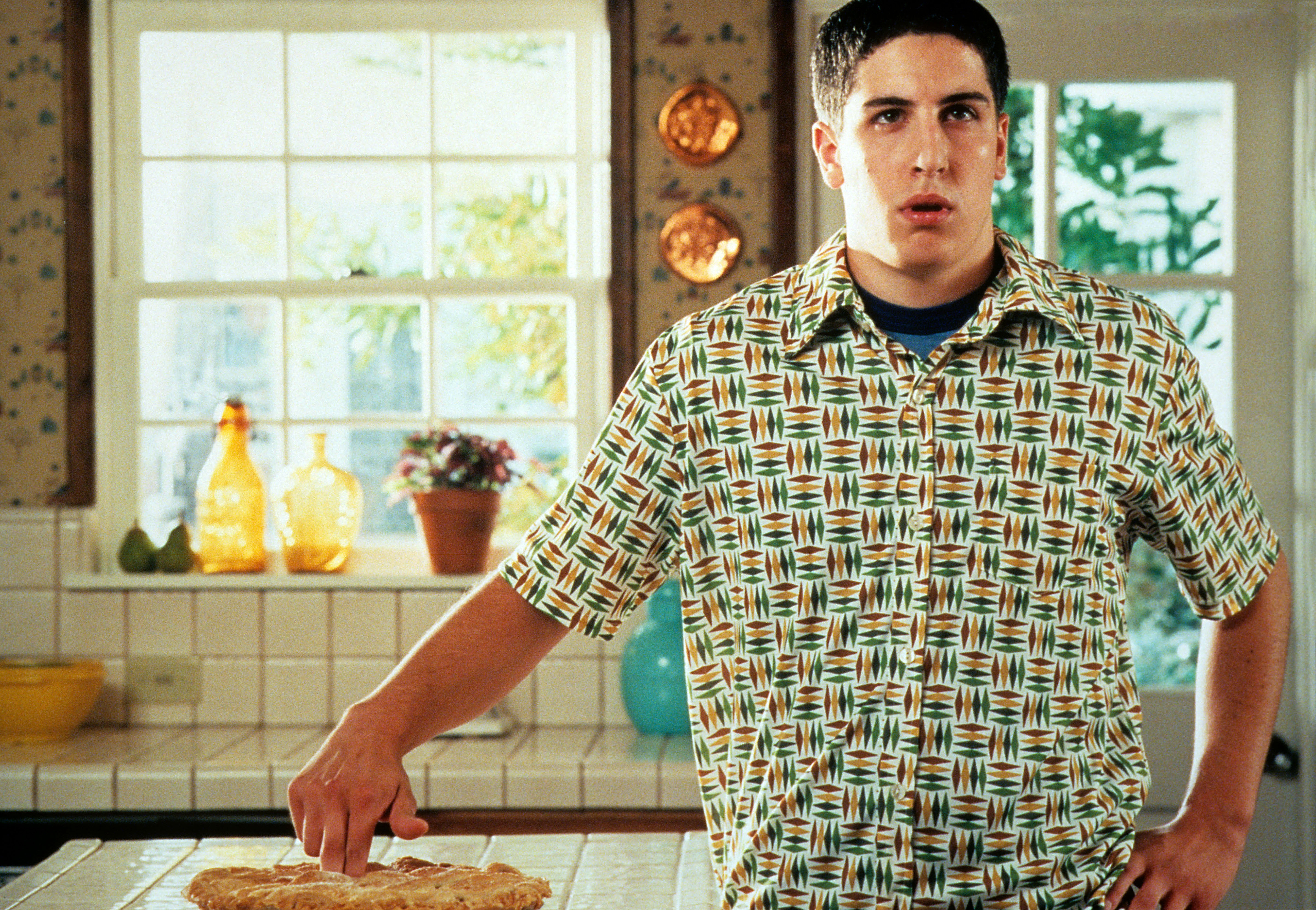 A person stands in a kitchen touching a pie with one finger, looking surprised, wearing a patterned shirt