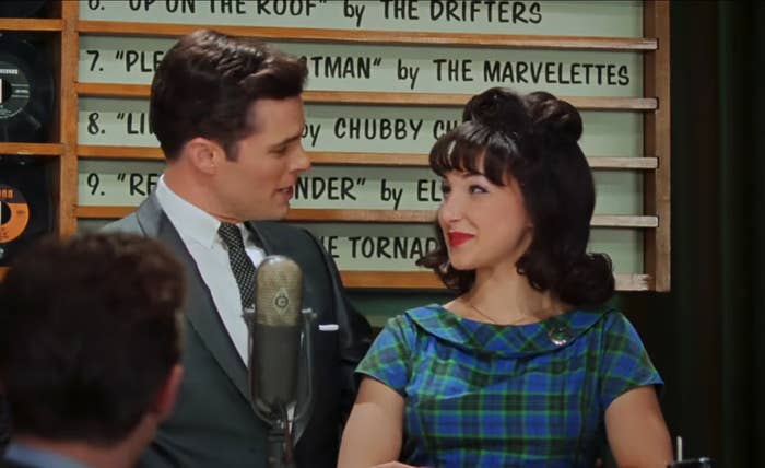 Man and woman in vintage attire engage in conversation near a retro microphone. Background features a vintage music chart