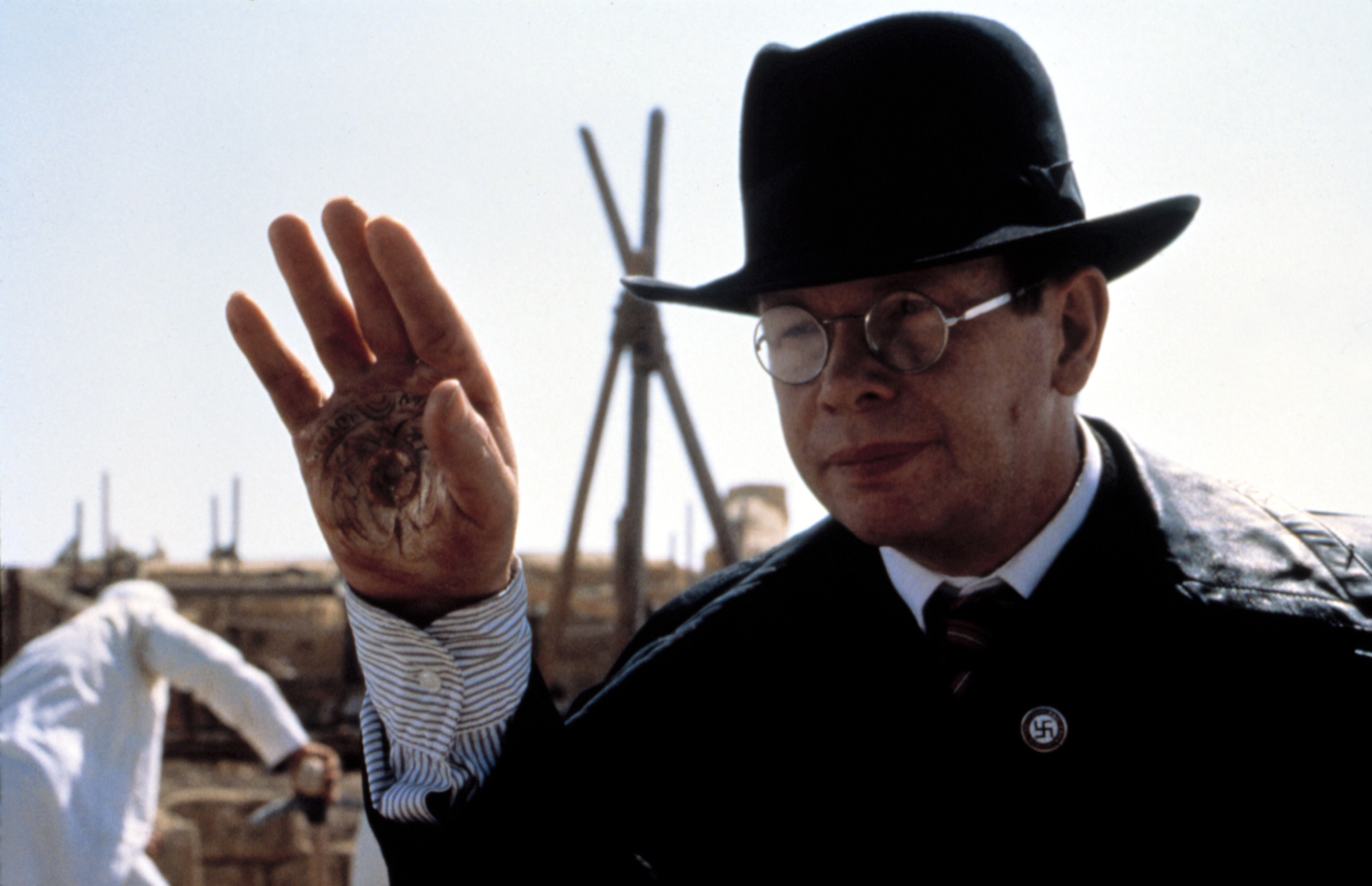 I don't know who this person is, but they are wearing a suit and hat, raising a hand with a burned symbol on the palm