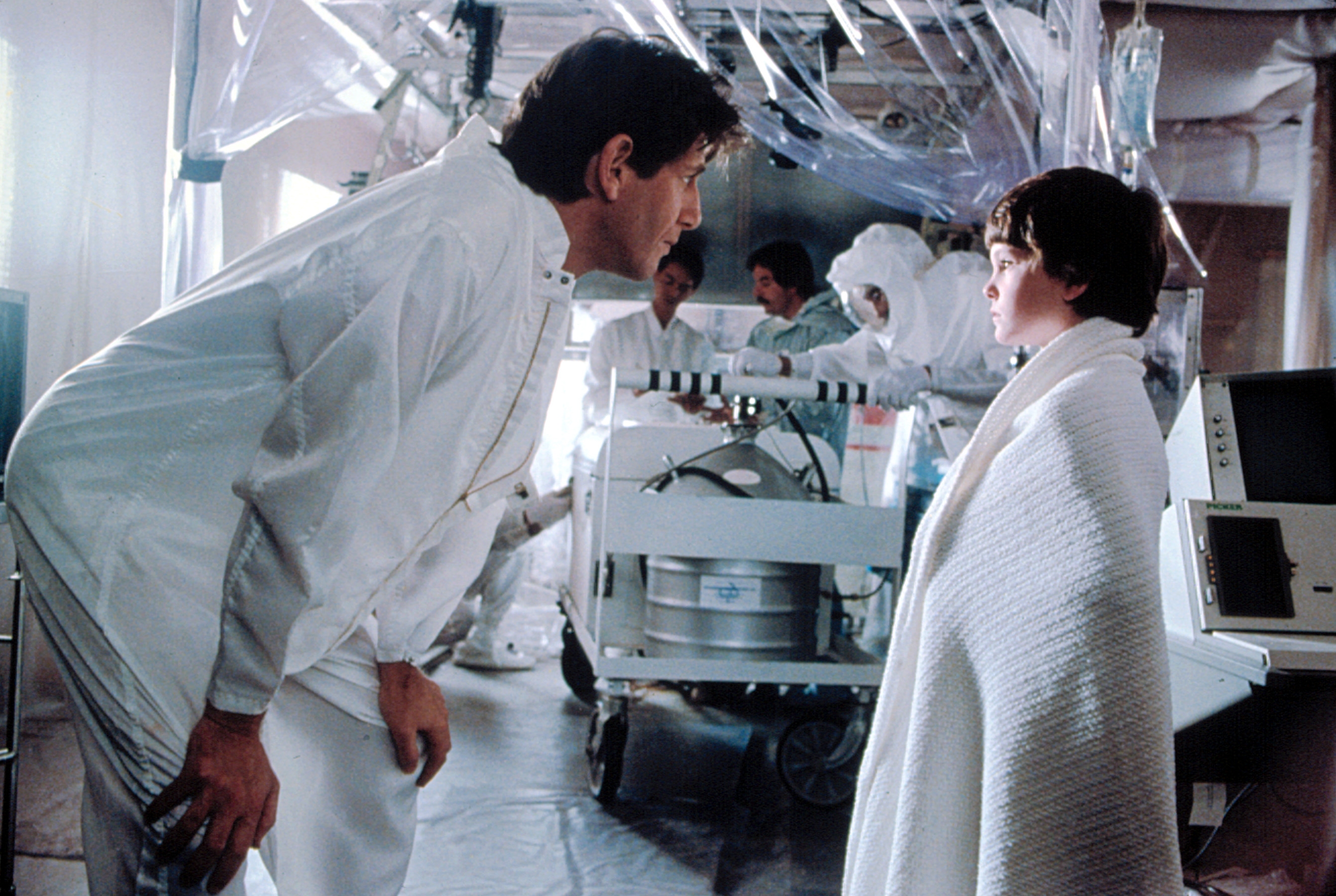 Man in a lab coat talks to a child wrapped in a blanket in a futuristic lab setting, with scientists working in the background