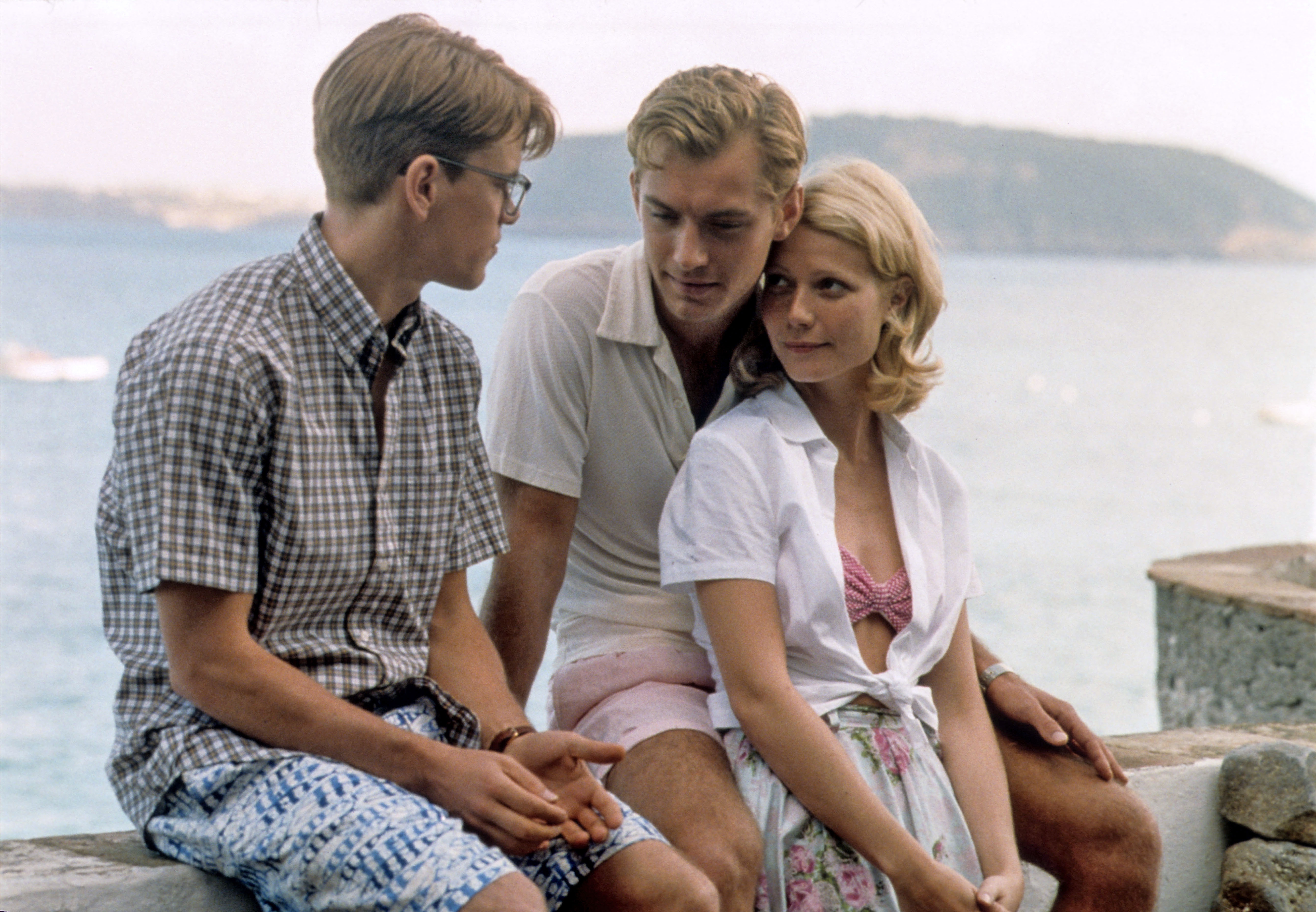 Three people sit outdoors by the water. They wear casual summer clothing, including shirts and shorts, and seem to be engaged in conversation