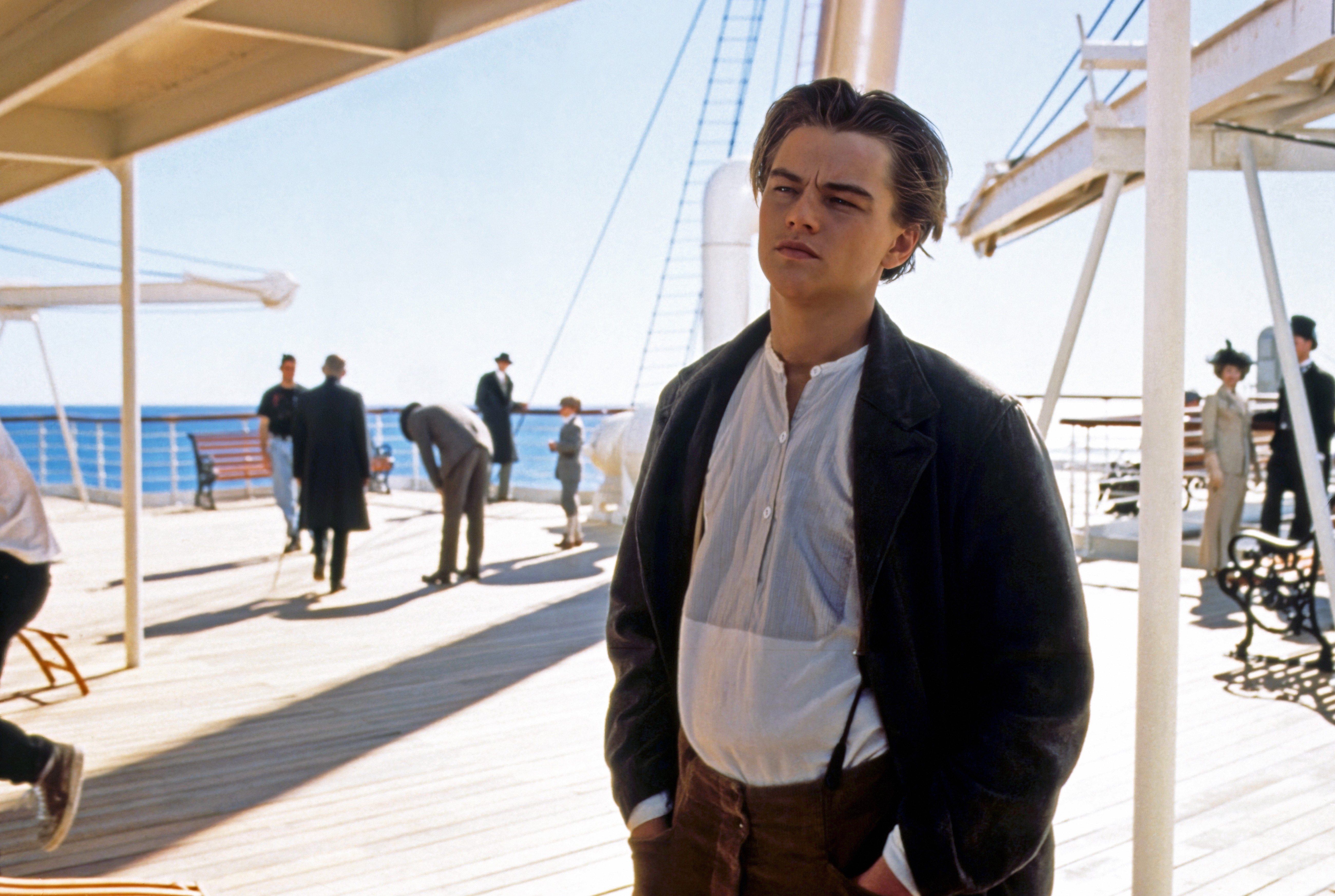 Person in period clothing stands pensively on a ship's deck with people in the background