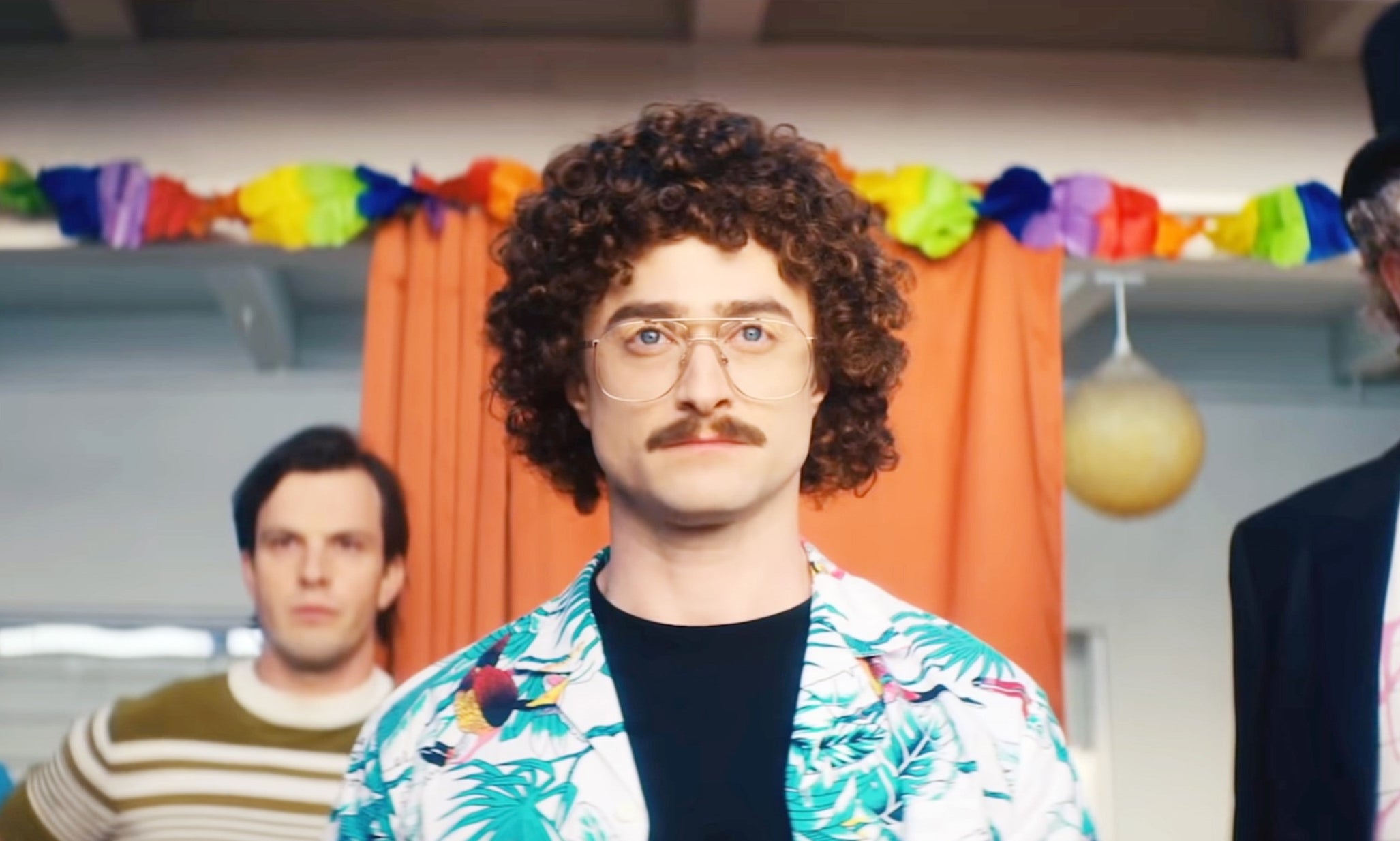 A group of four people stand together in a room, one in a floral shirt and glasses, another in a top hat, likely from a TV or movie scene
