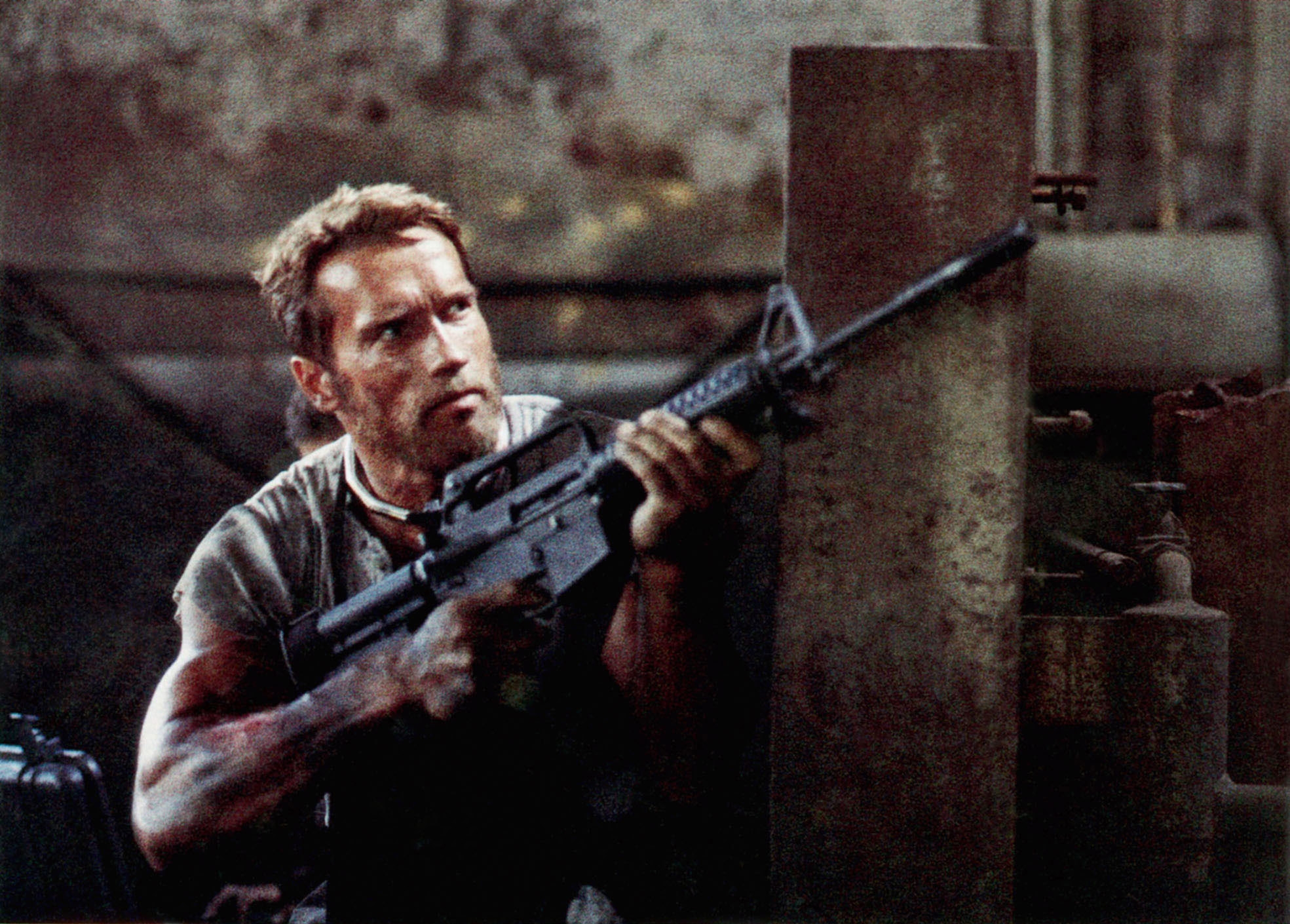 A muscular man with short hair, holding a rifle, crouches behind cover in an action scene from a movie