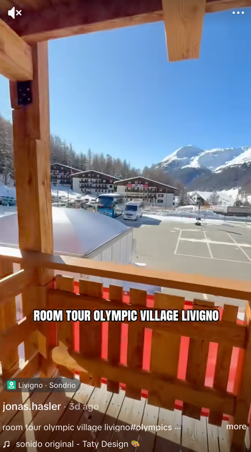 View from a woody  balcony overlooking buildings and snow-covered mountains successful  Livigno's Olympic Village. Text: "Room circuit  Olympic Village Livigno."