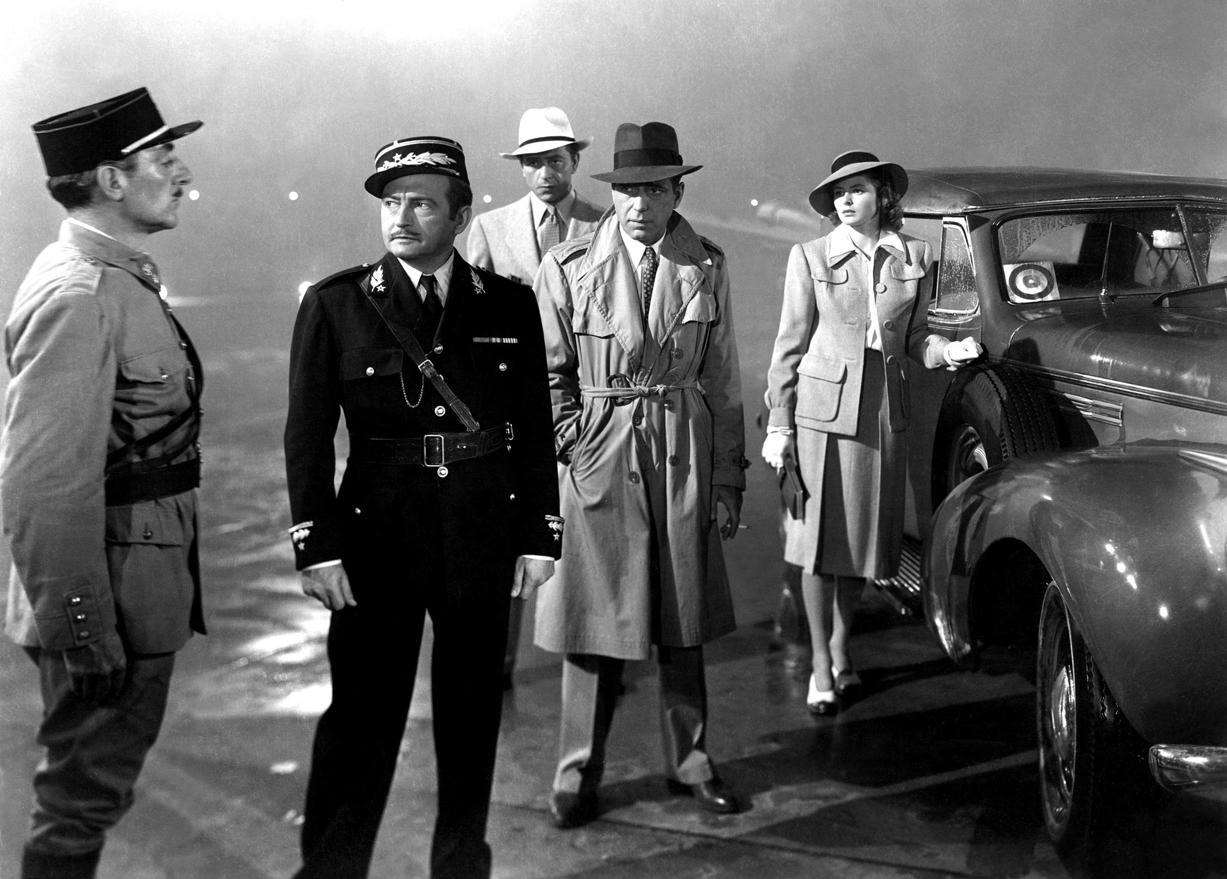 Group of five people in 1940s attire stand by a classic car on a misty street; one wears a military uniform, others in trench coats and suits