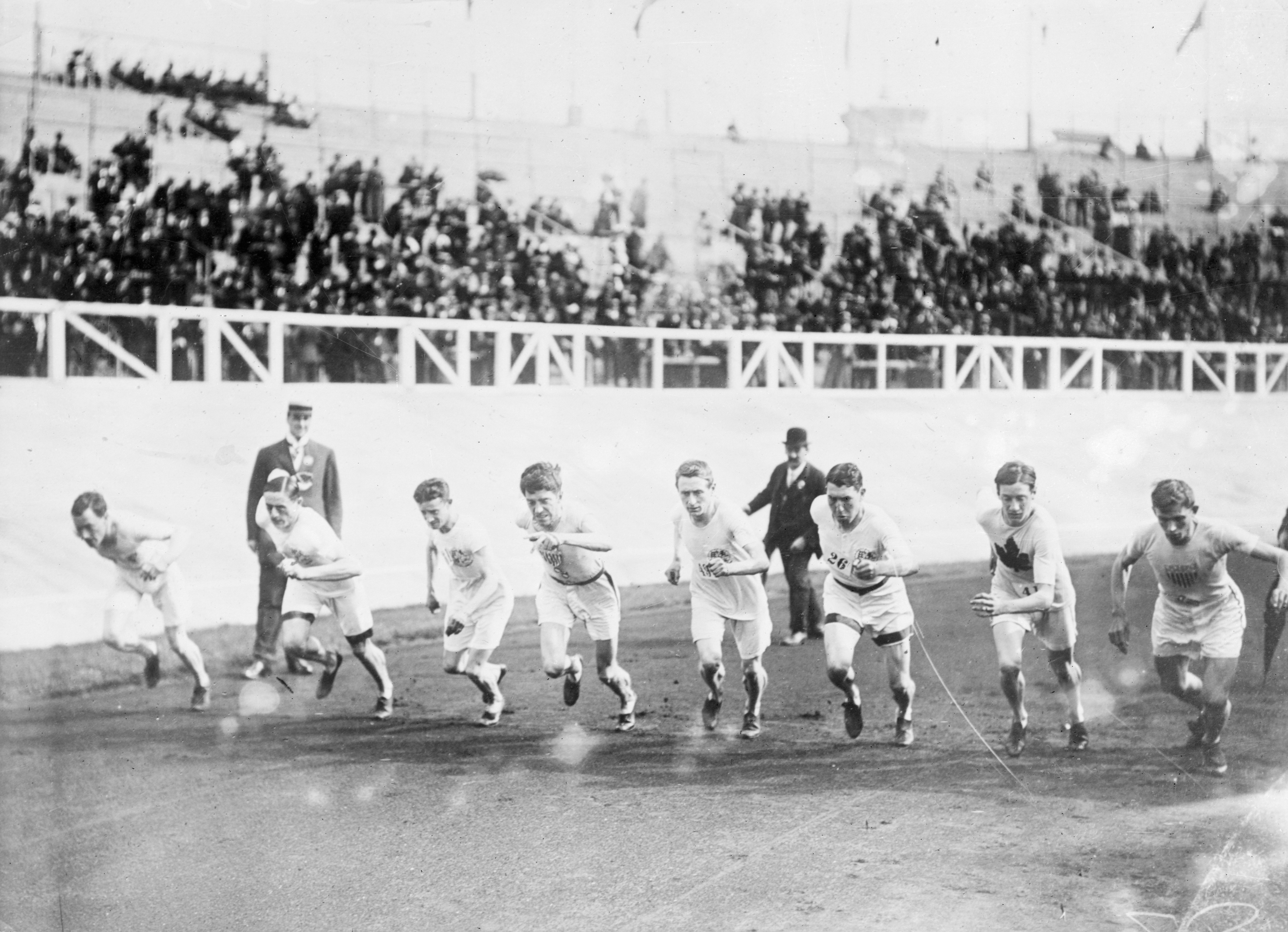 Historic photo of athletes in running gear starting a race on a track, with spectators in the background. Early 20th-century sports event