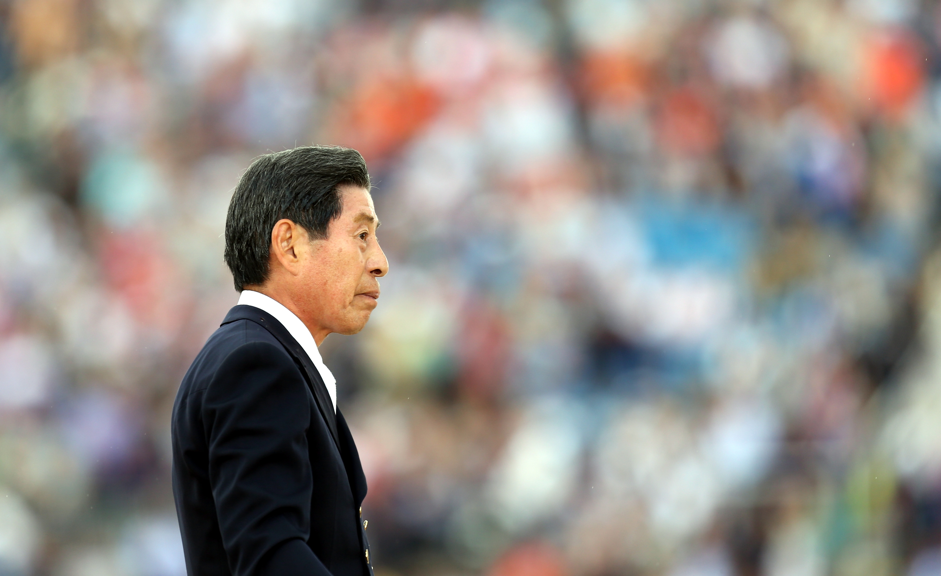 Person in formal suit walking outdoors at an event with a blurred crowd in the background