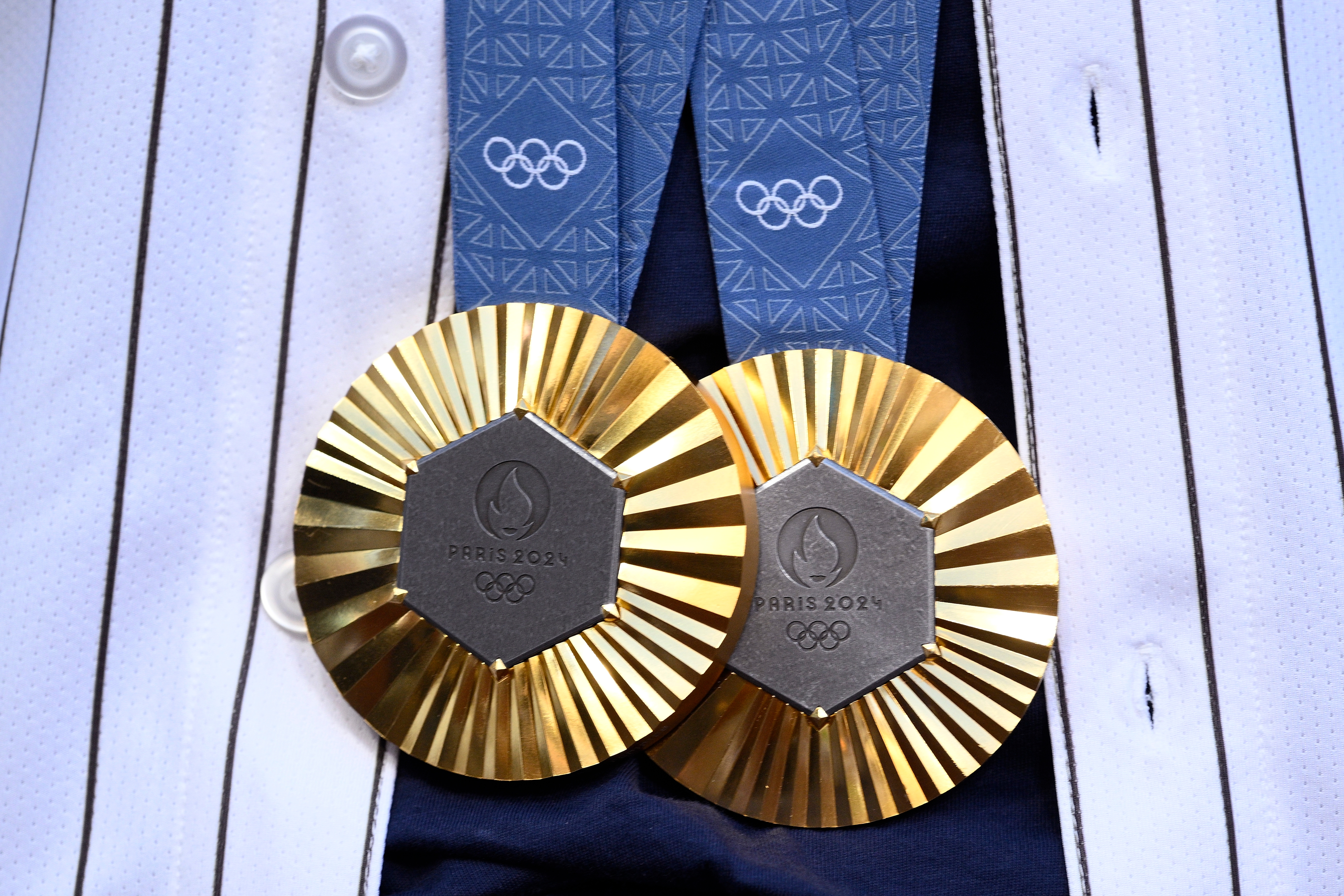 Two gold Olympic medals, Paris 2024, with a geometric pattern, hanging on a striped fabric background