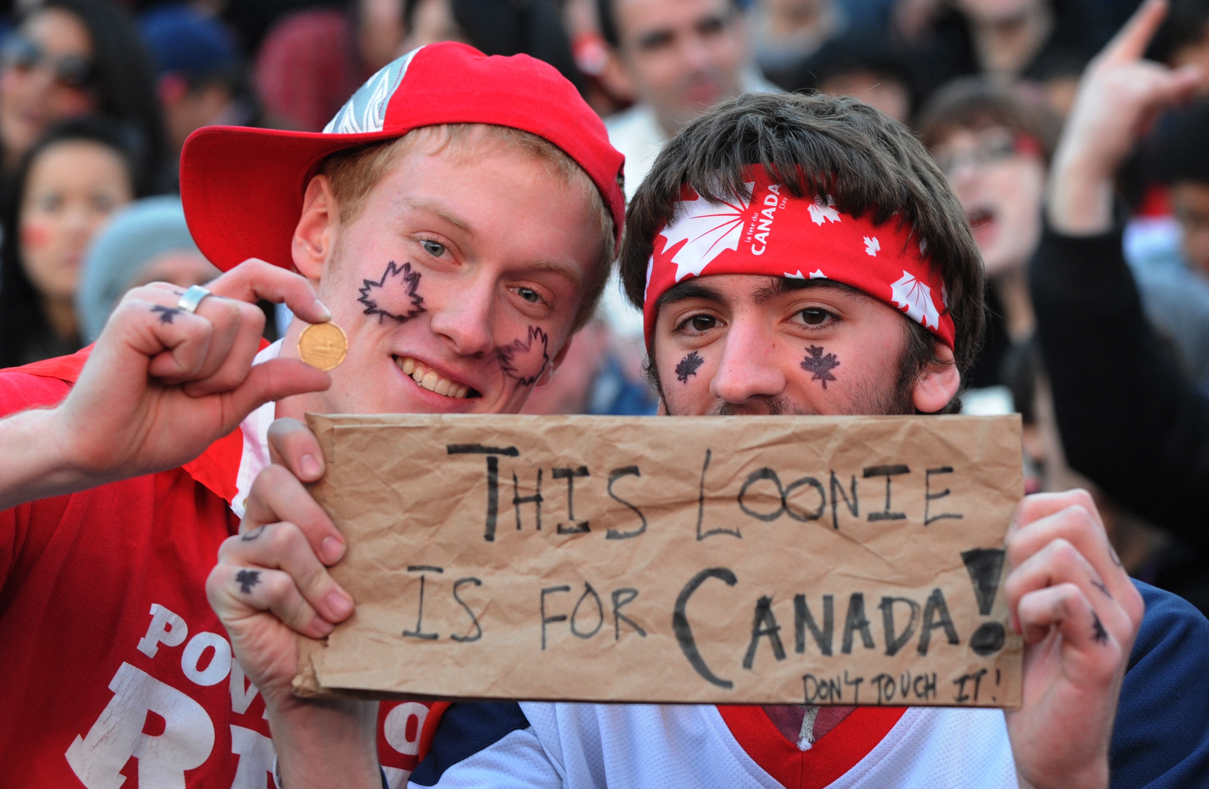 Two people with face paint and Canada-themed accessories hold a sign that reads, "This loonie is for Canada! Don't touch it!" One shows a coin