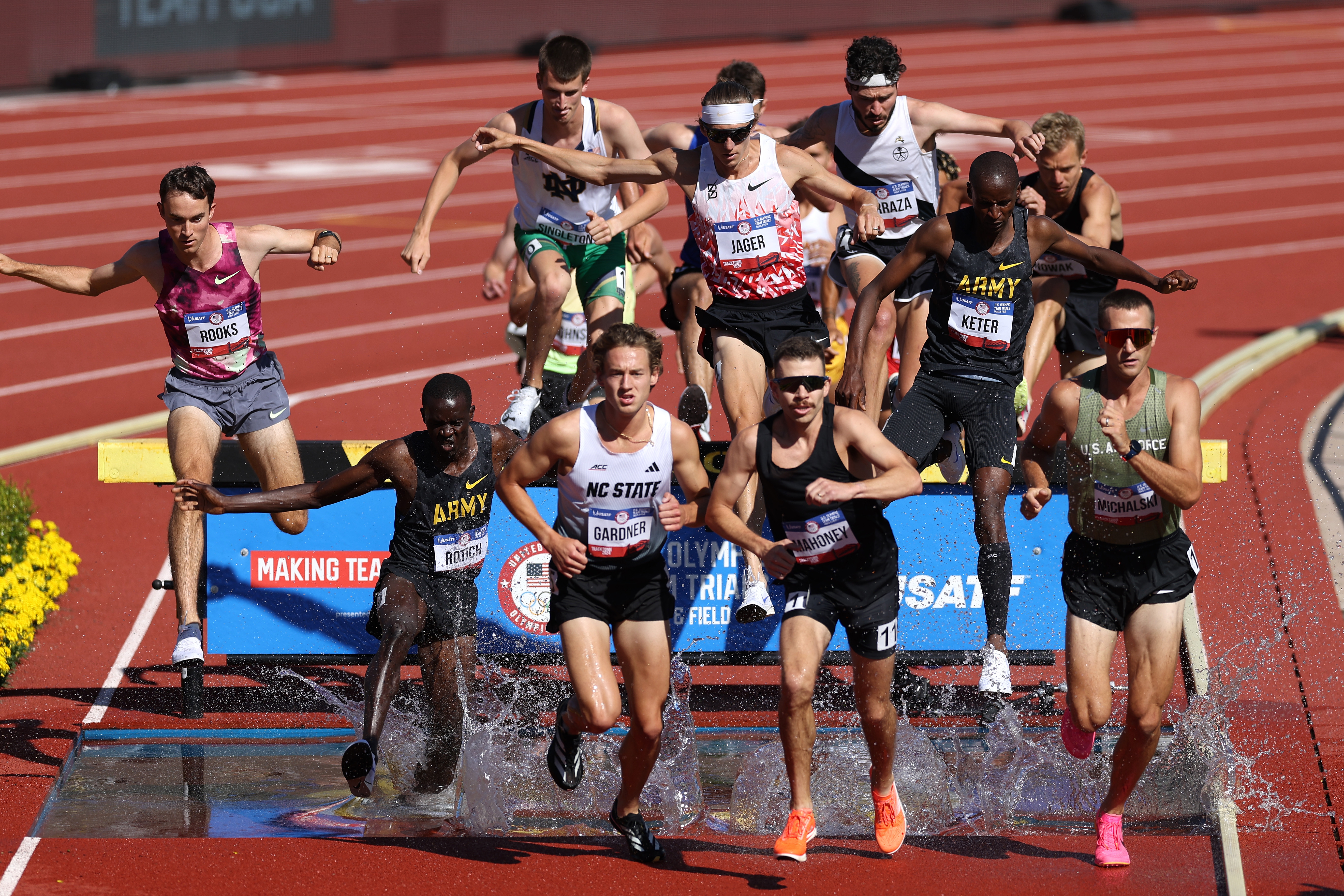 Athletes in a track and field steeplechase event leap over water-filled barriers on a sunny day, displaying focus and agility