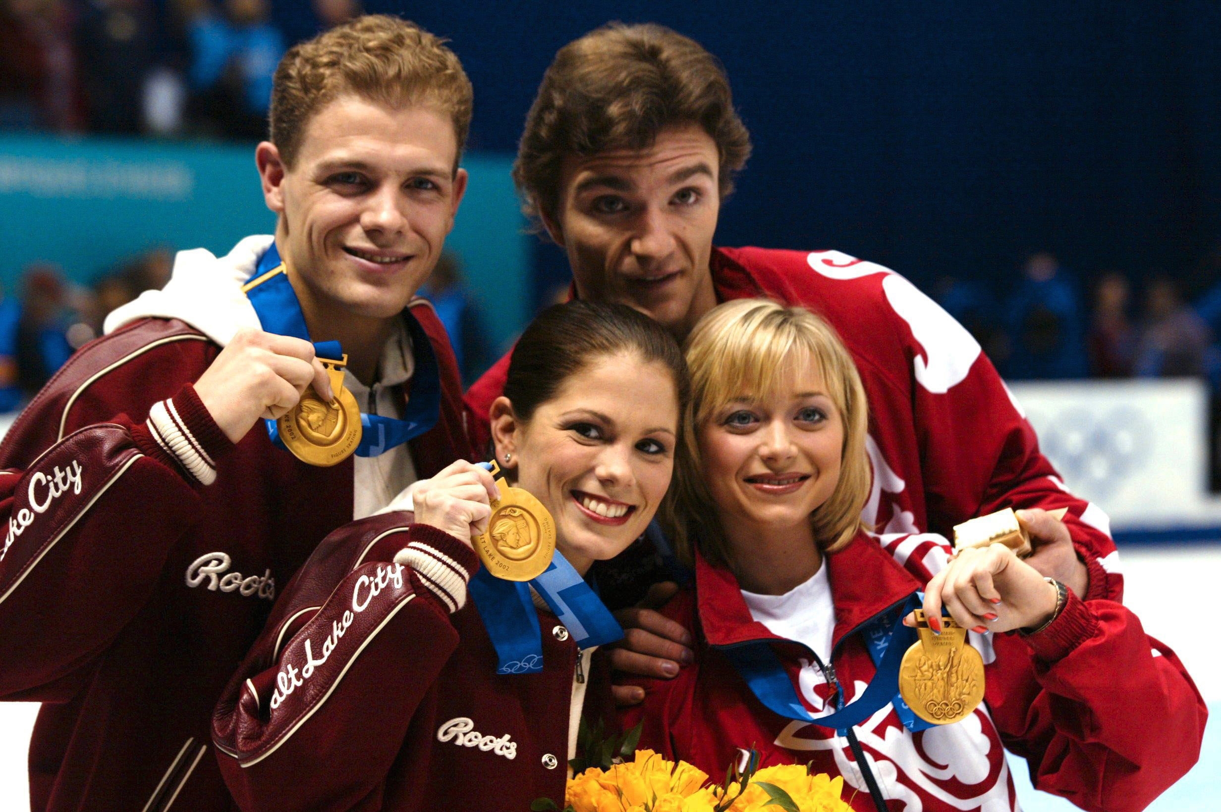 Four figure skaters proudly display their gold medals and bouquets, smiling broadly, wearing matching jackets at a skating event