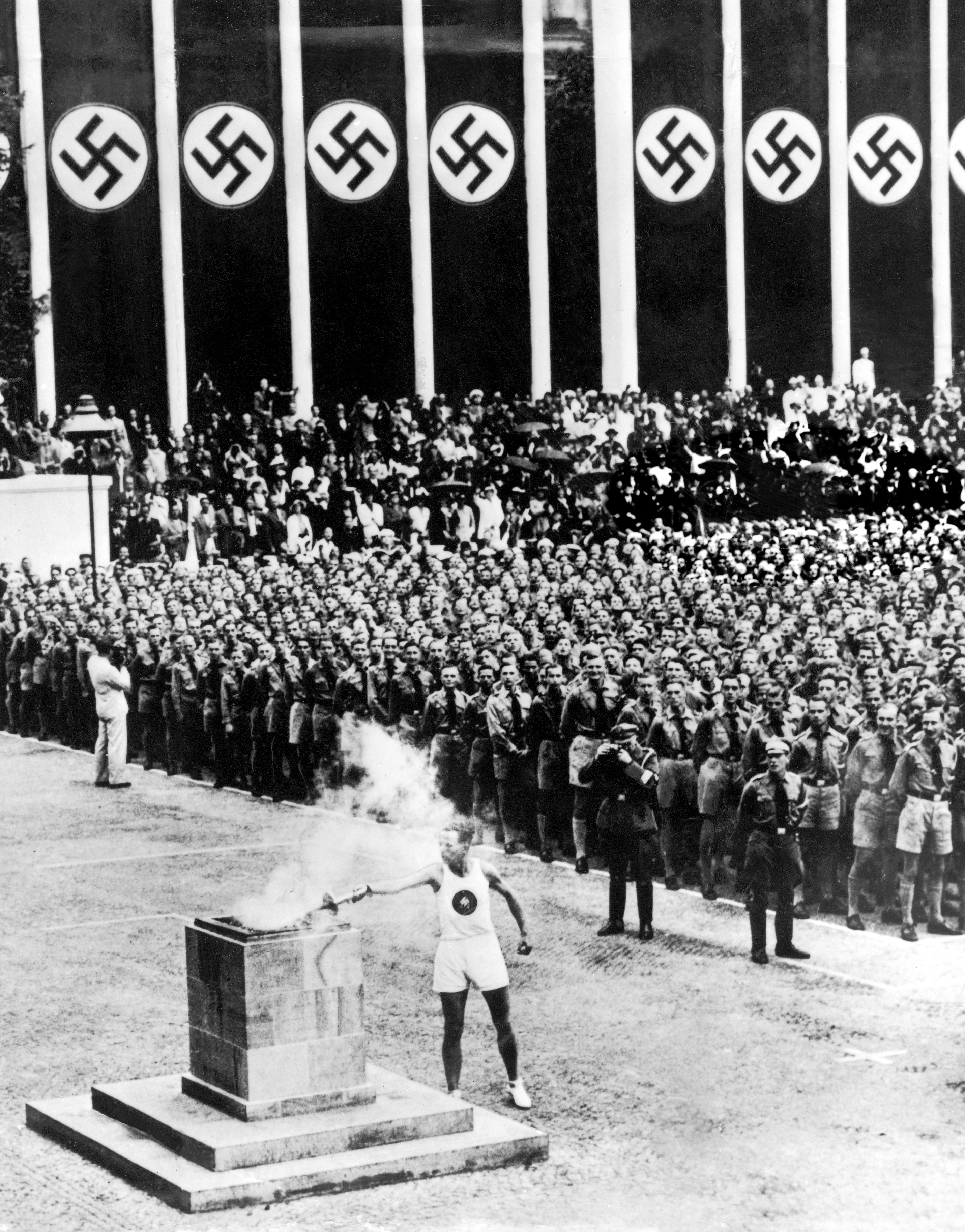 Athlete lights Olympic torch at 1936 Berlin Games opening, with Nazi symbols and uniformed soldiers in the background