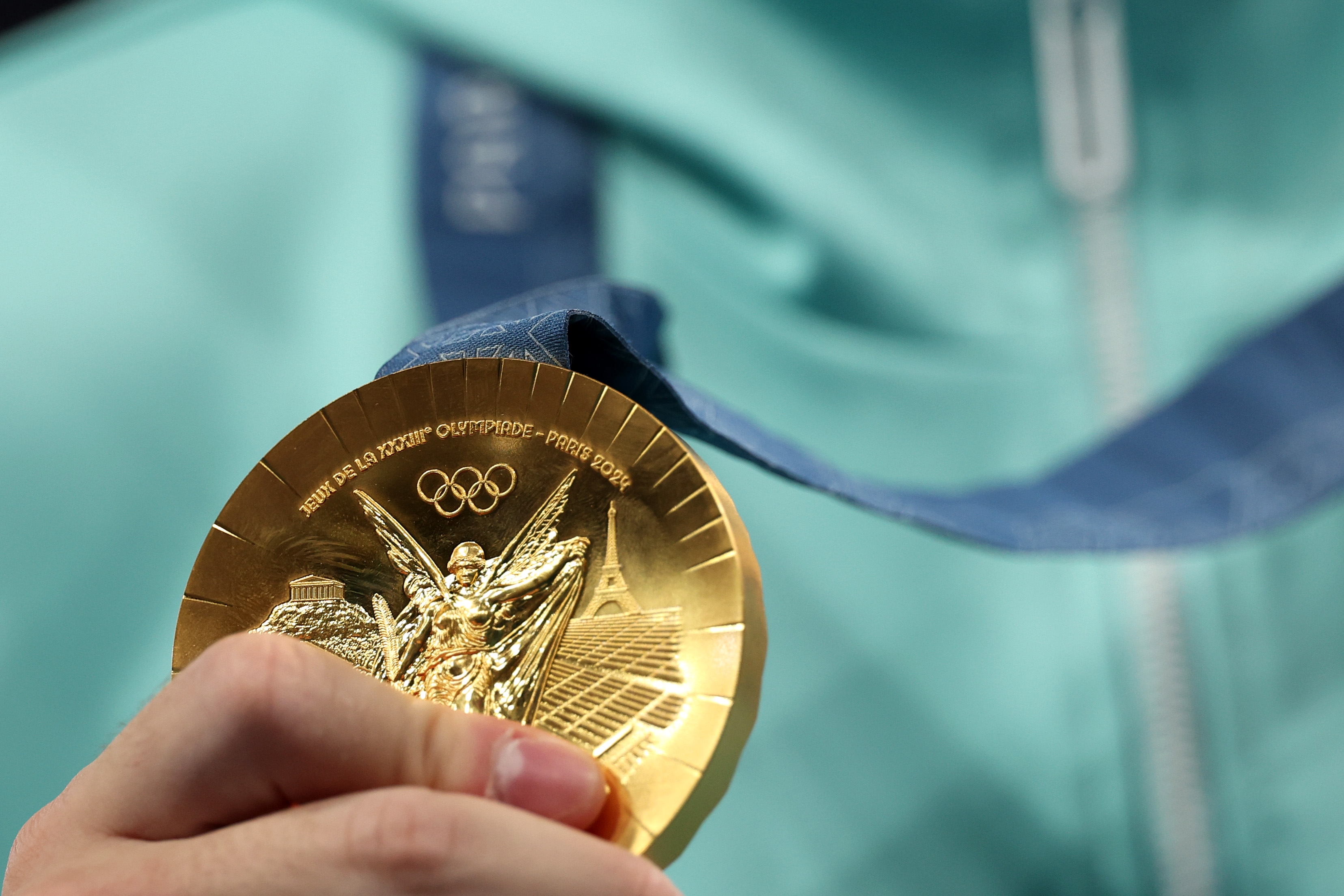 Hand holding an Olympic gold medal with embossed details and a blue ribbon visible