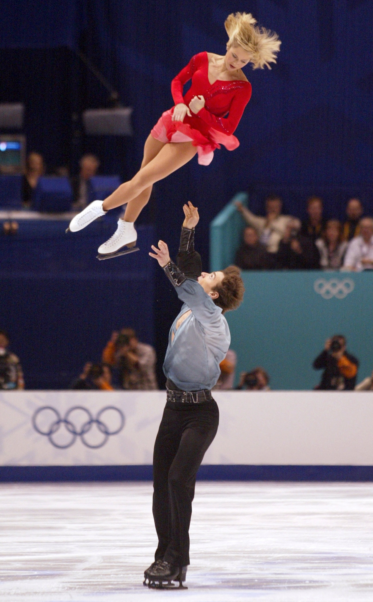 Figure skating pair performing an acrobatic lift on ice during an Olympic event, with the female skater in a dress and the male in a shirt and pants