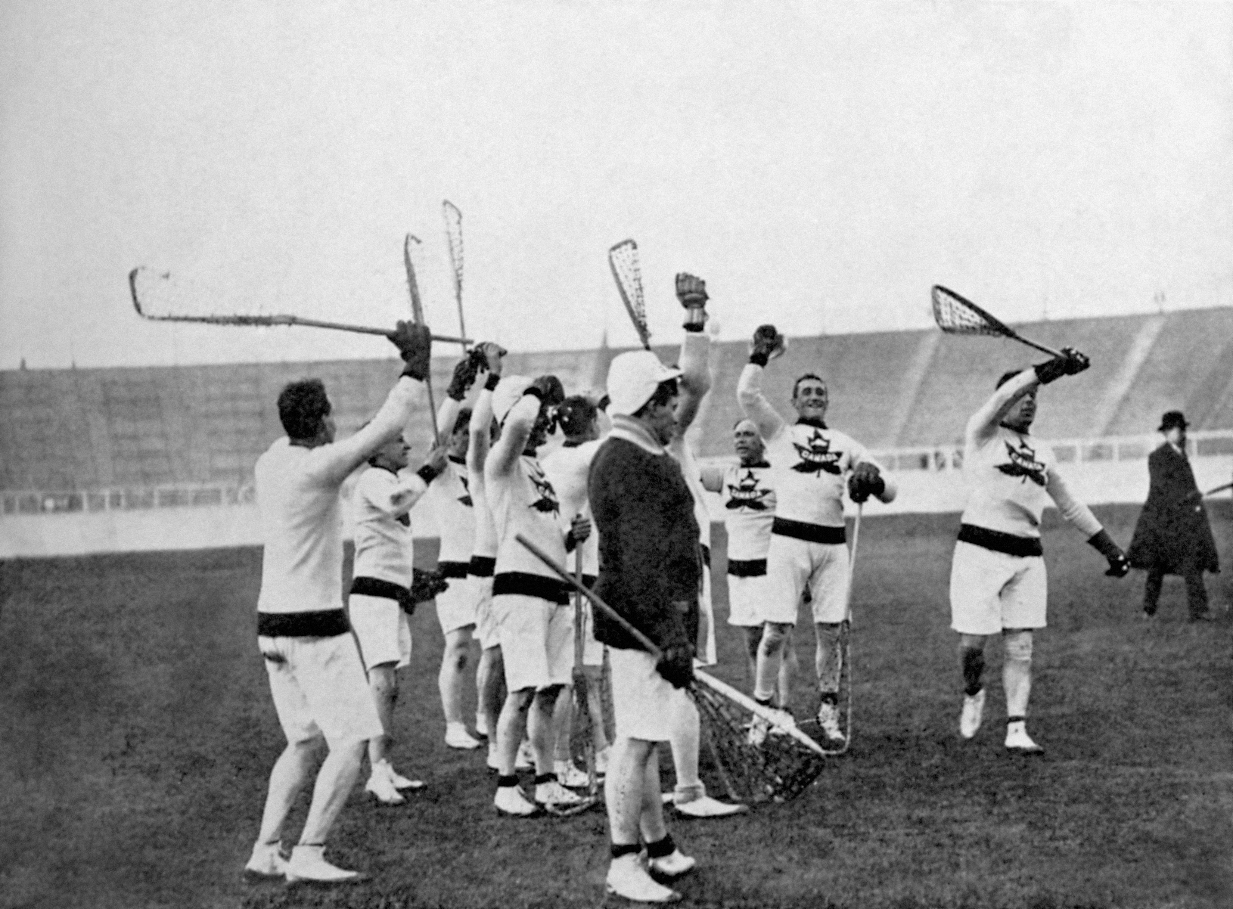 Early 20th-century lacrosse players in uniform celebrate with raised sticks on a field. Stadium stands visible in the background