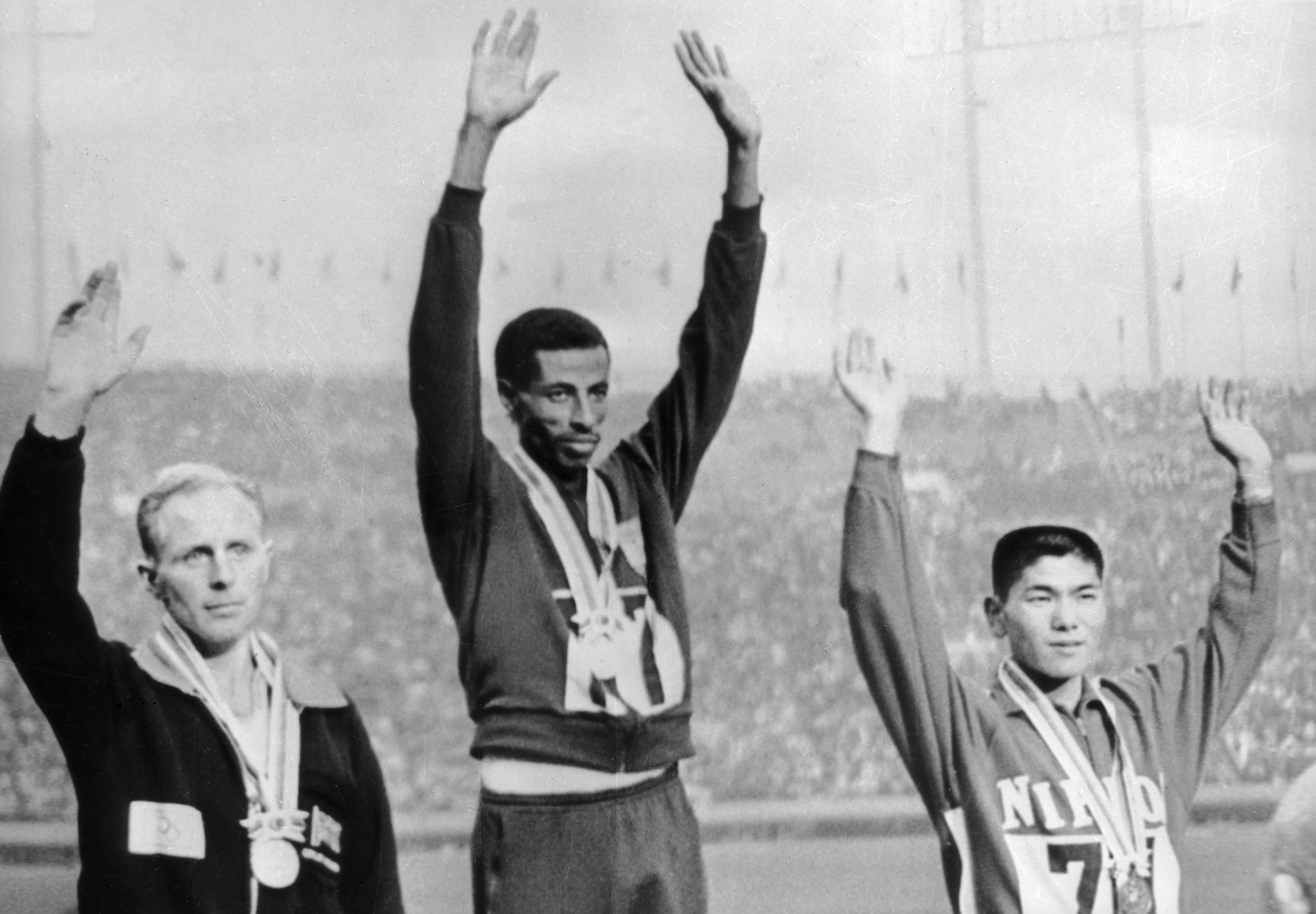 Three athletes on a podium raise their arms. The central athlete holds the gold medal, flanked by silver and bronze medalists