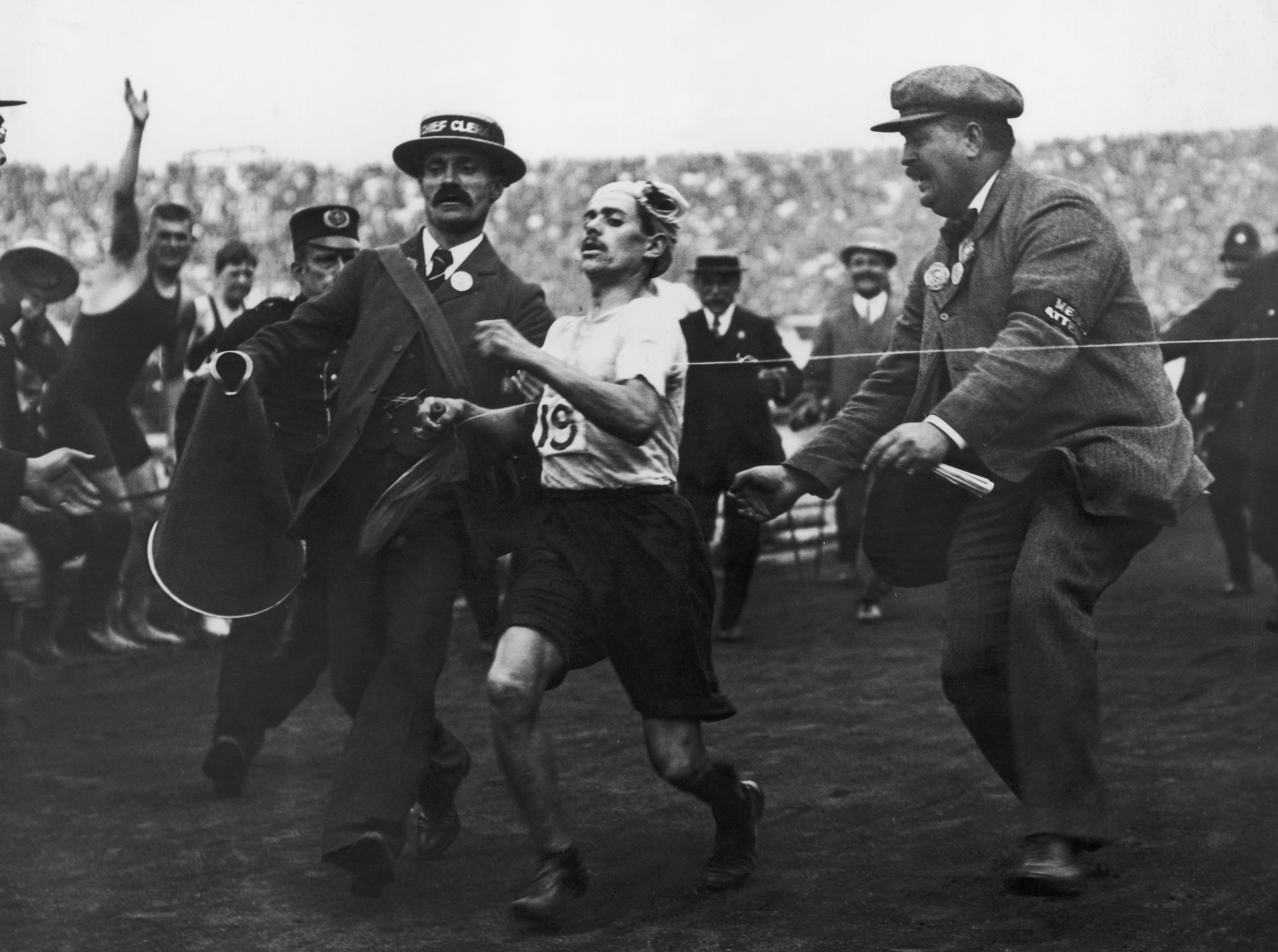 Early 1900s marathon scene; fatigued runner assisted to finish line by two men in hats; crowd in background